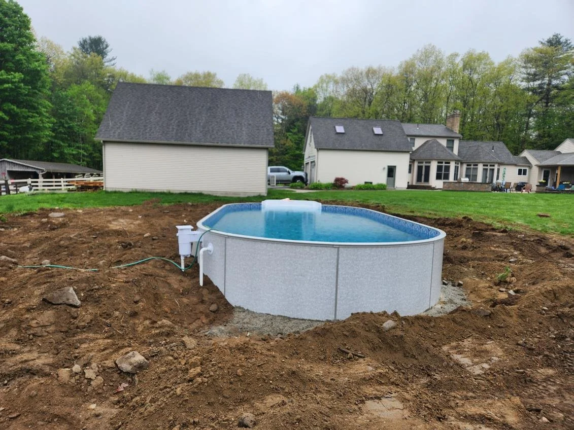 Above ground swimming pool with a blue interior and white exterior, partially filled with water, on a patch of dirt in a backyard, with houses and trees in the background.