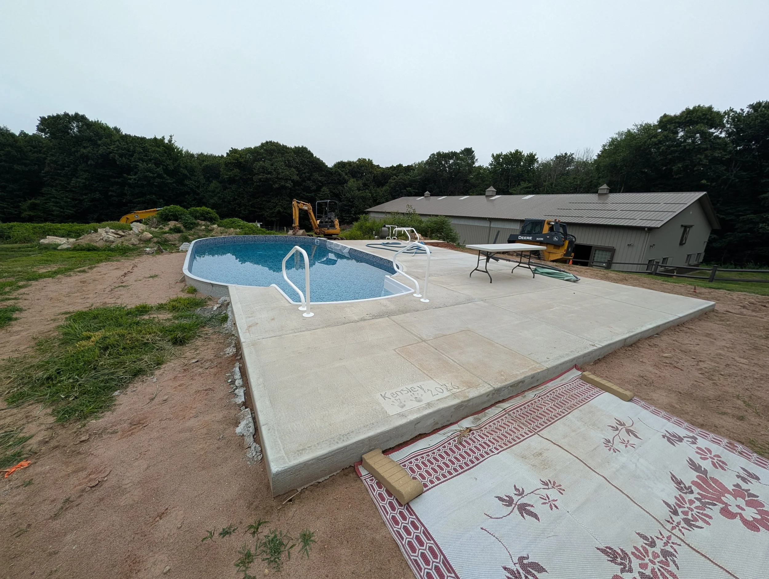 Construction site near a pool with building materials, machinery, and a house in the background.