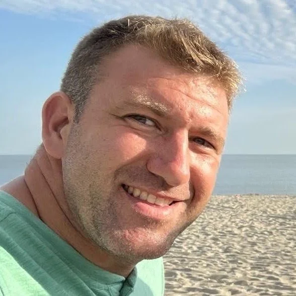 A man smiling at the camera on a beach with sand and the ocean in the background, under a partly cloudy sky.