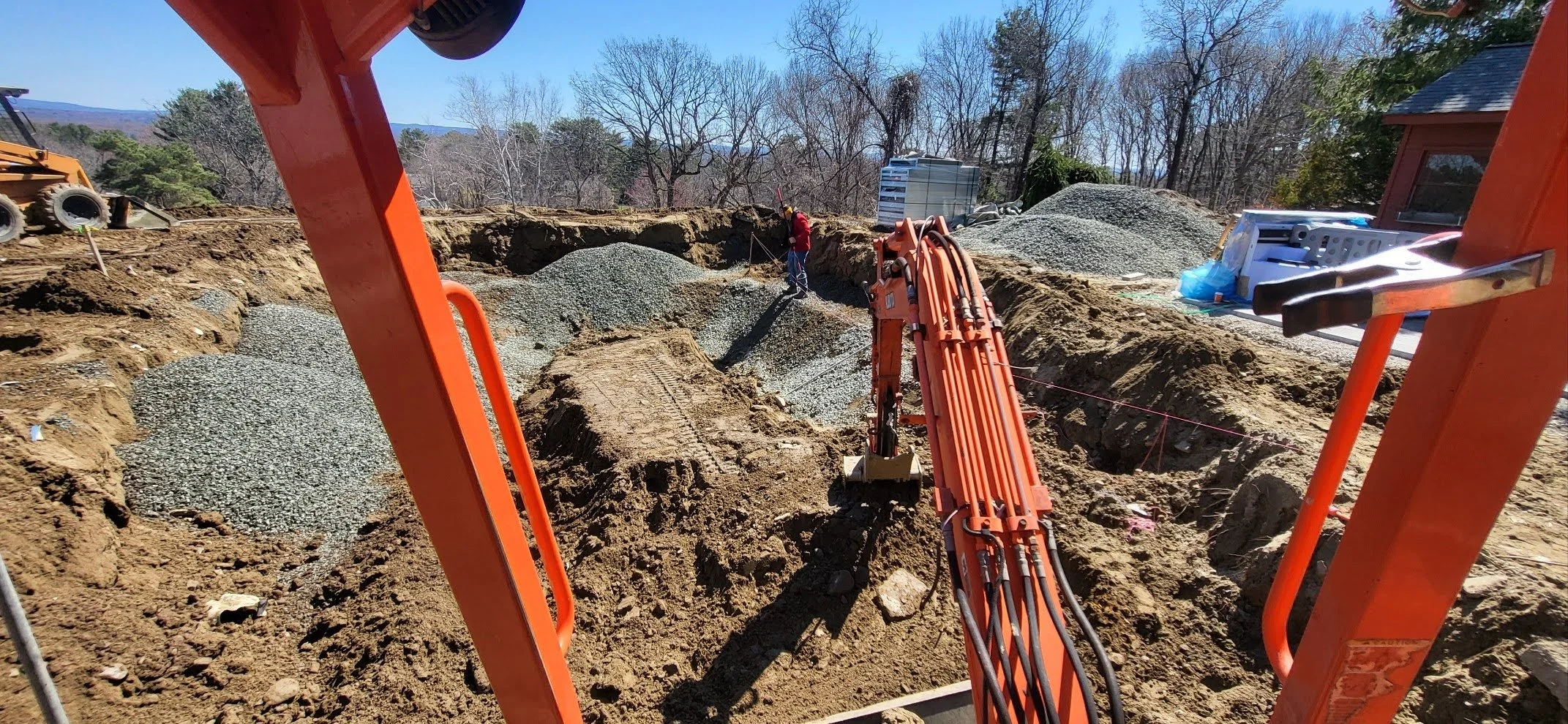 Construction site with heavy machinery and a worker, digging a trench with gravel and dirt piles, surrounded by trees and clear sky.