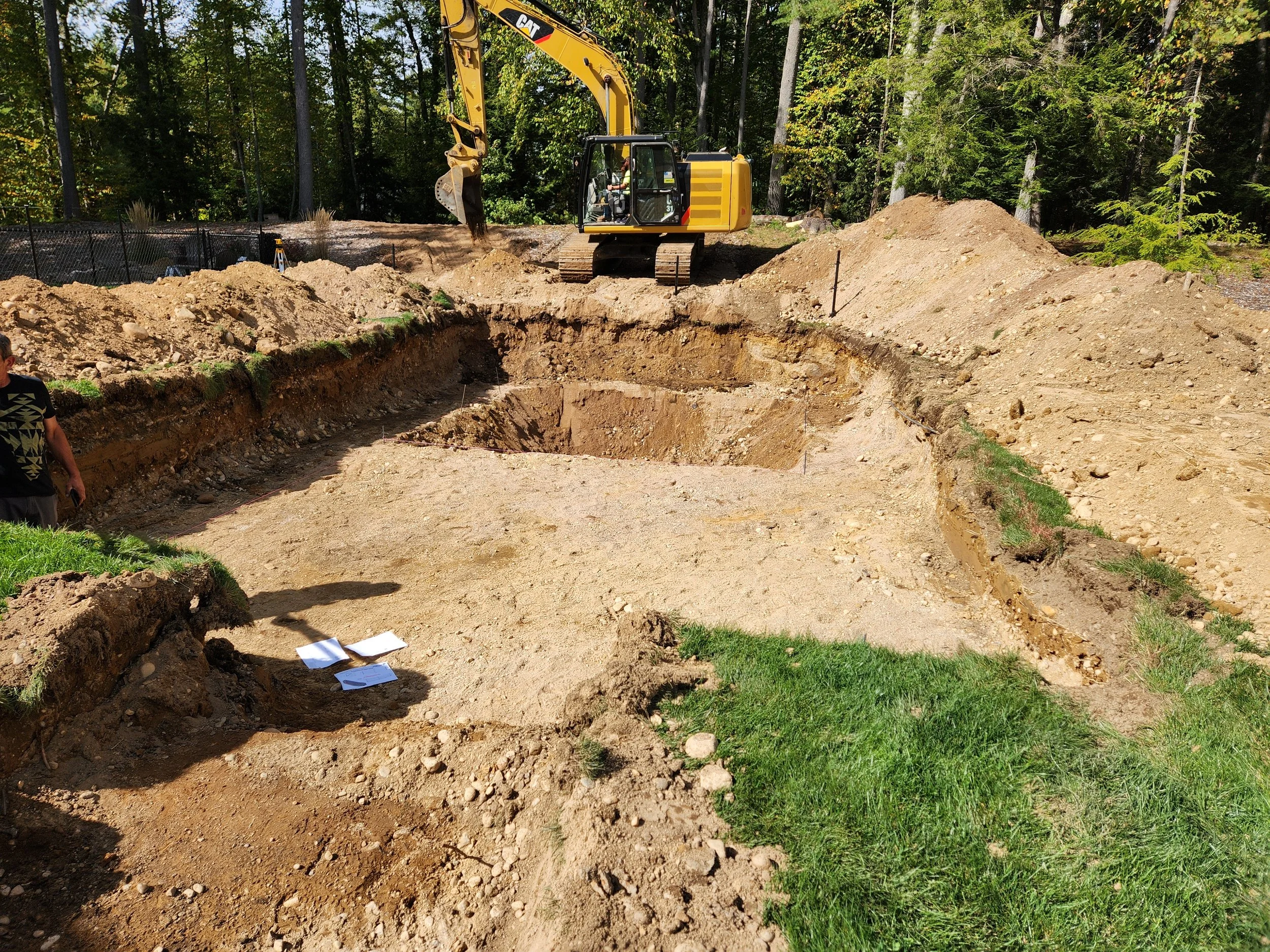 Construction site with a yellow excavator digging a hole in the ground surrounded by dirt mounds, with trees in the background.