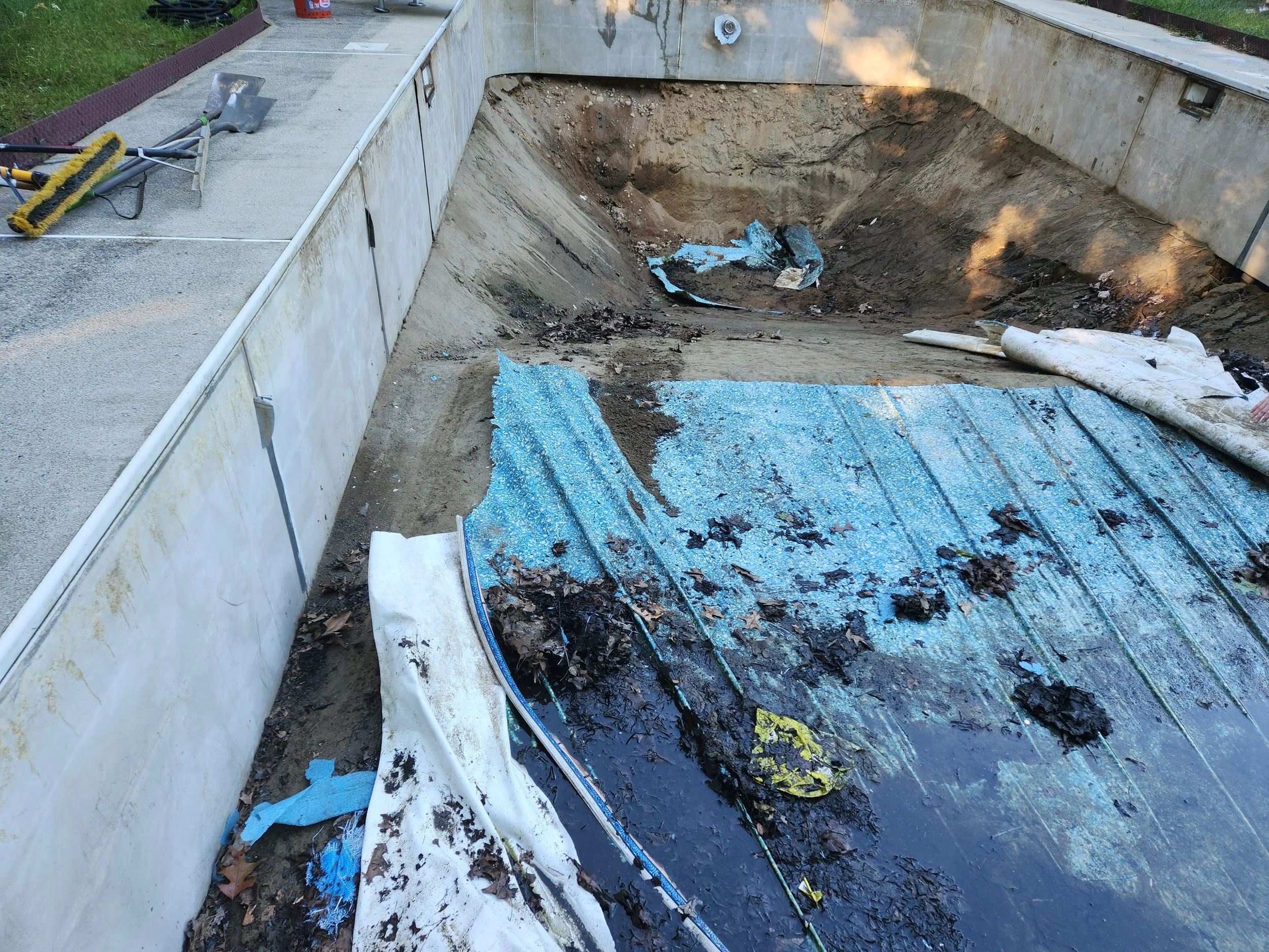 An empty swimming pool under construction with the liner removed, revealing dirt and debris inside, along with tools and materials on the edge.