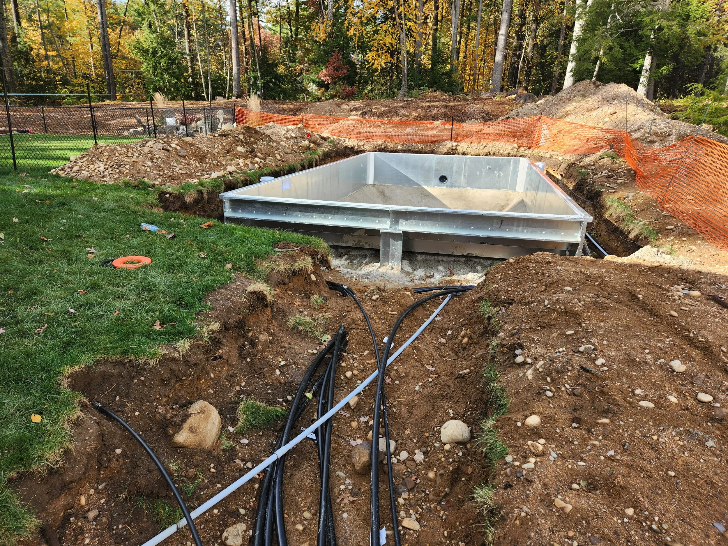 Construction site with an in-ground pool frame, black and orange safety fencing, dirt piles, and trees in the background.