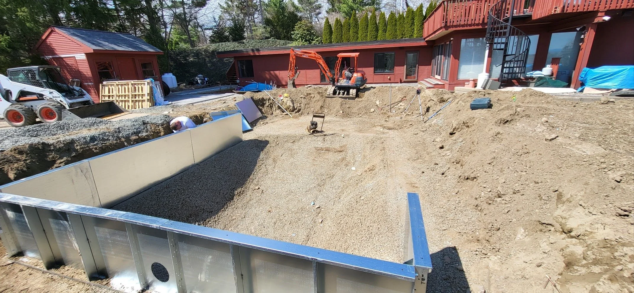 A construction site in the backyard of a red house with a spiral staircase, featuring an excavator, construction materials, and a partially built foundation surrounded by dirt.