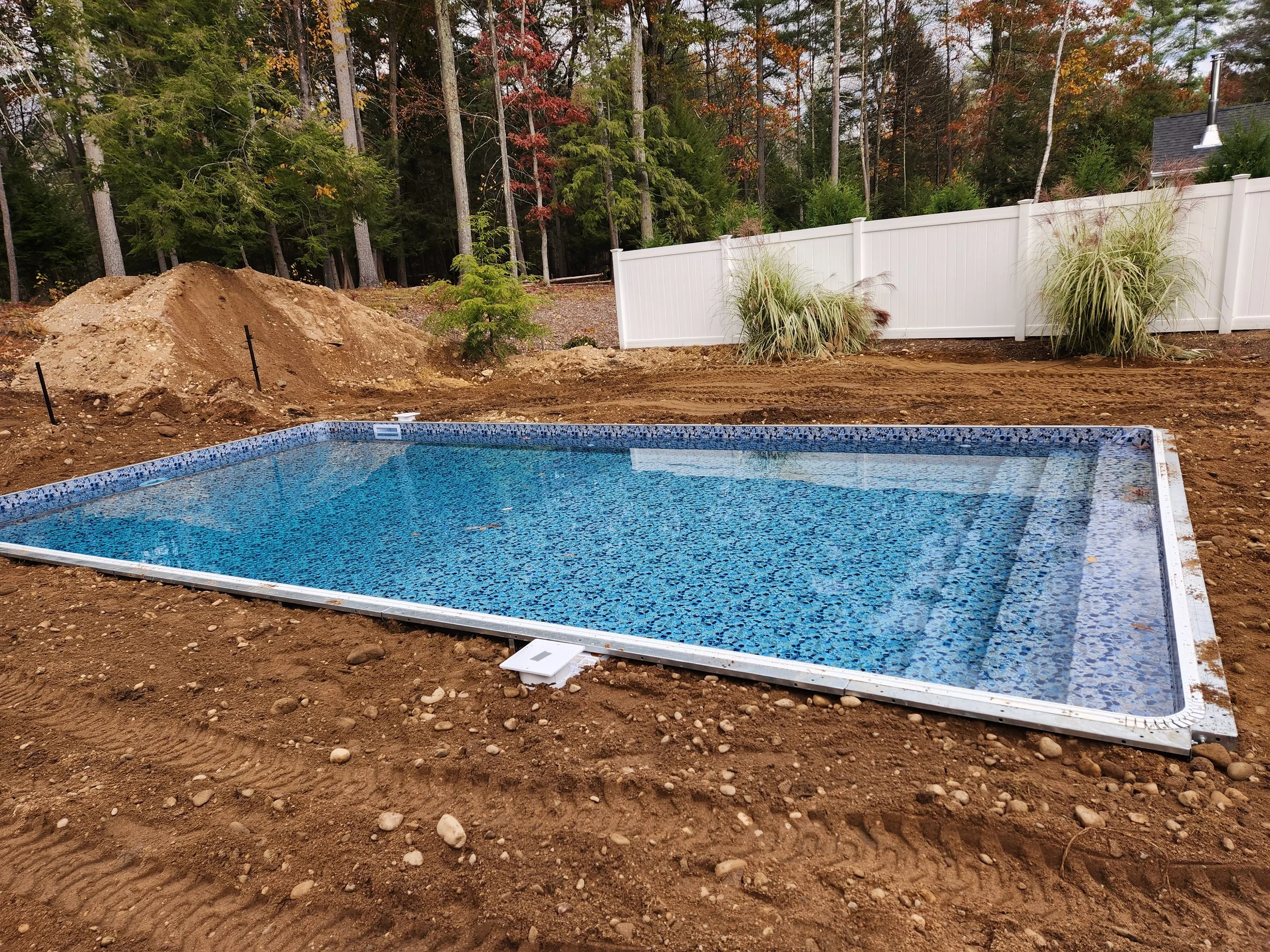 A backyard scene under construction featuring a blue tiled swimming pool surrounded by dirt, a white privacy fence, and trees with fall foliage in the background.