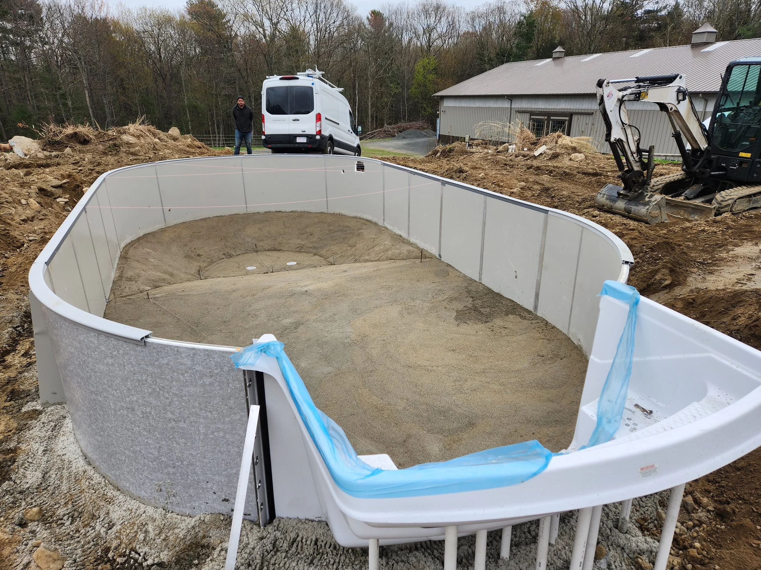 Construction site with a large circular foundation, a man standing near a white van, a small excavator, and a house in the background.