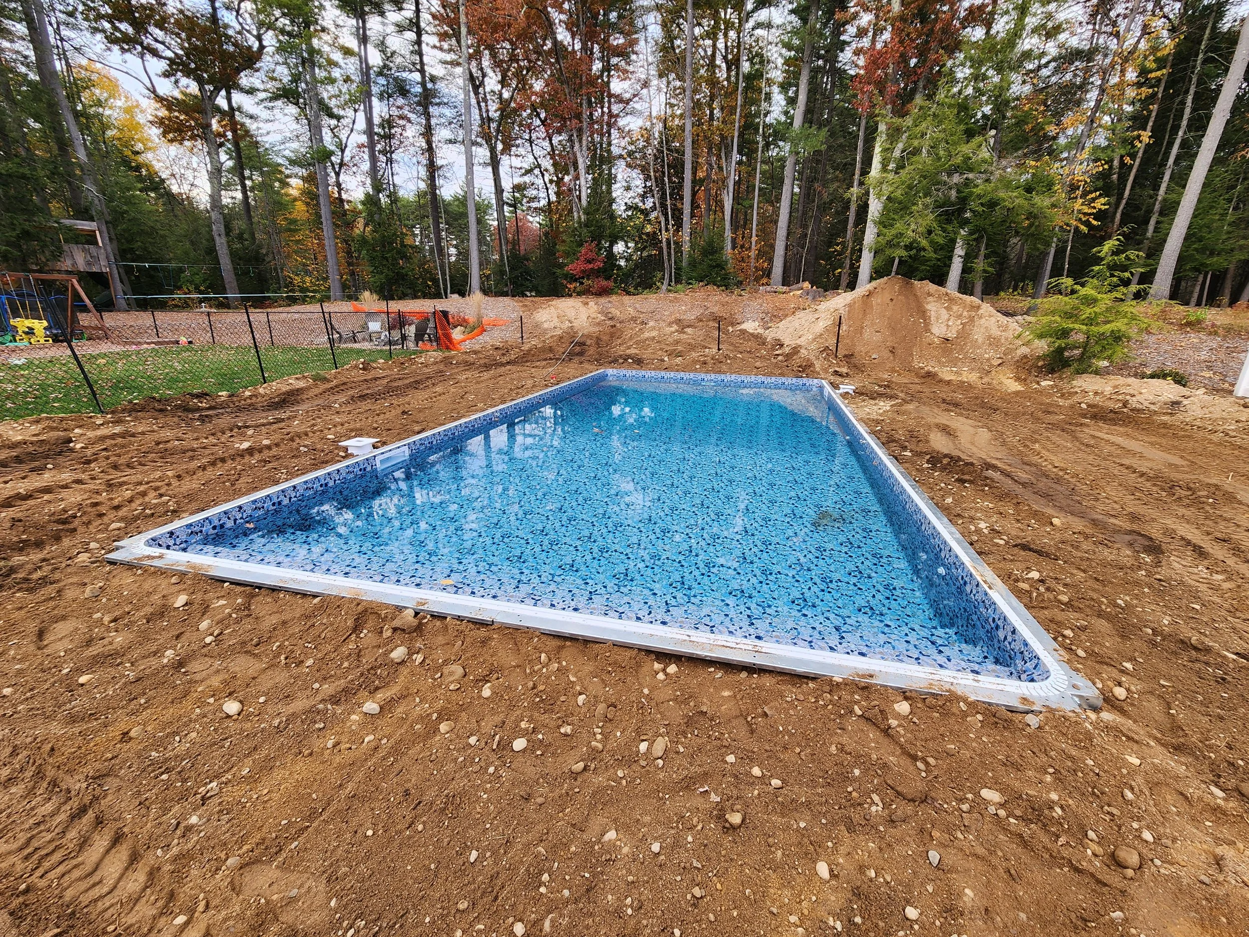 Construction site with a partially completed in-ground swimming pool, dirt and soil around, surrounded by trees with autumn foliage.