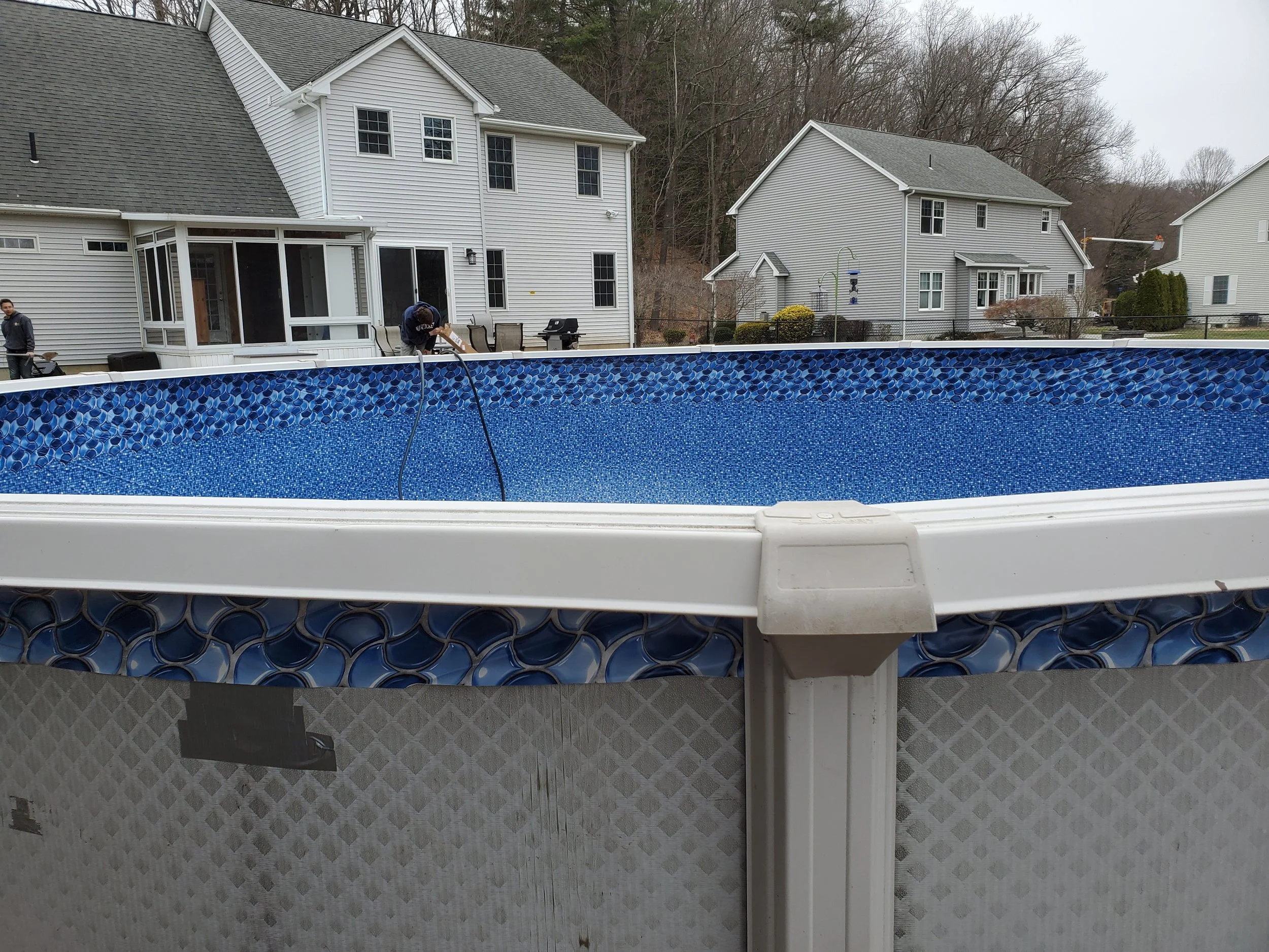 A view of a backyard with a deep blue swimming pool with mosaic tile border, white house, and neighboring houses in the background, on a cloudy day.