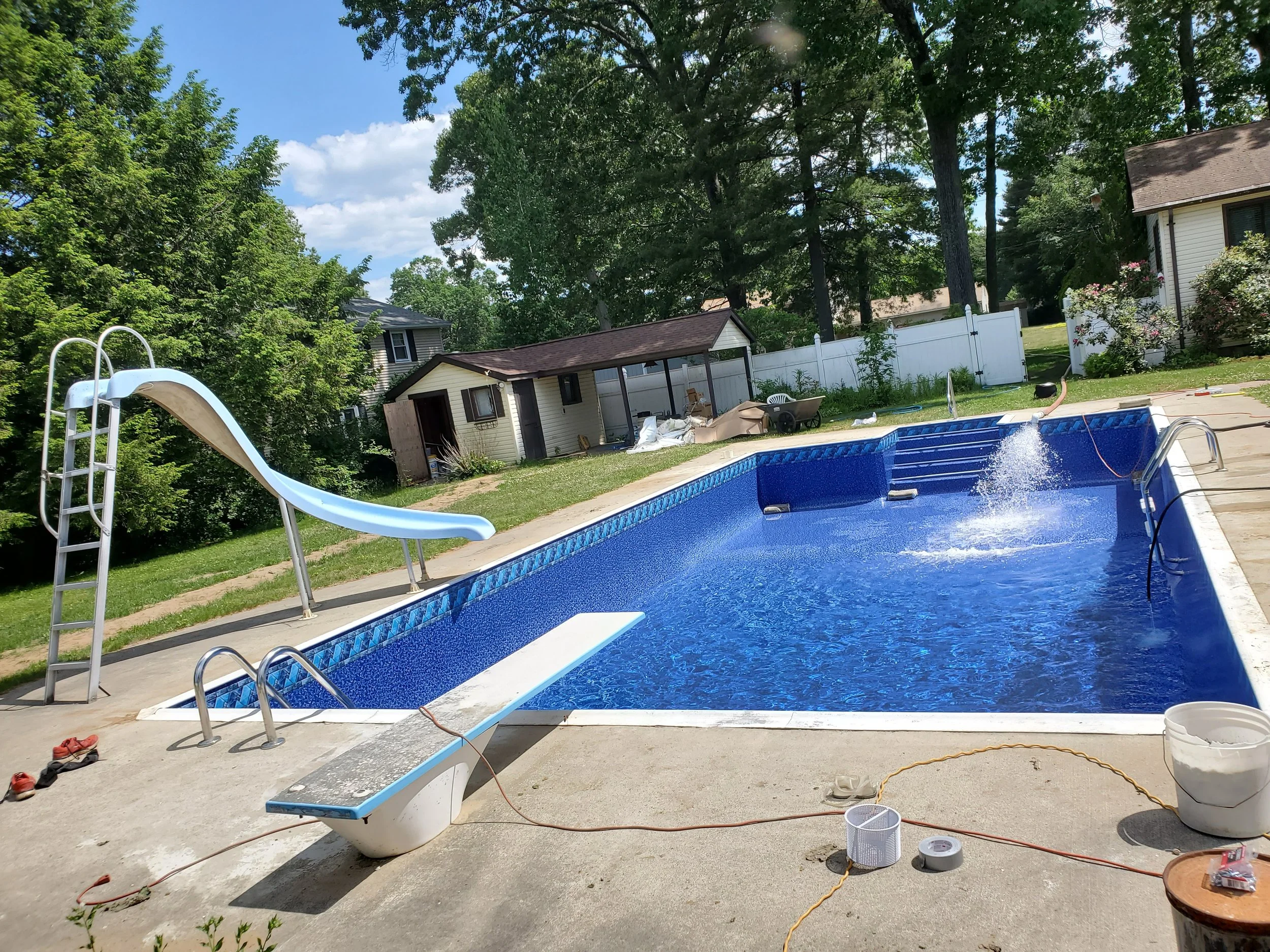 A backyard swimming pool under construction with a slide, surrounded by lawn and trees, and various tools and materials scattered around.
