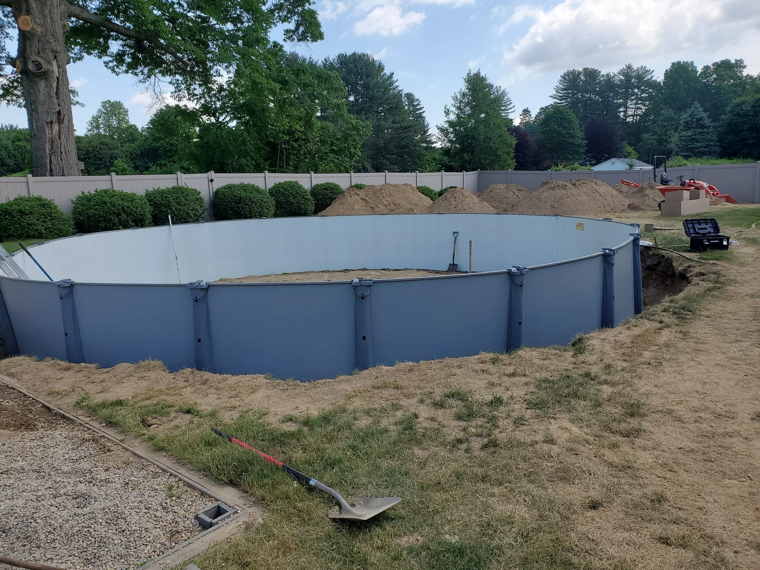 Backyard with a pool under construction, surrounded by a white fence, with piles of dirt, shrubs, trees, and construction tools and equipment.