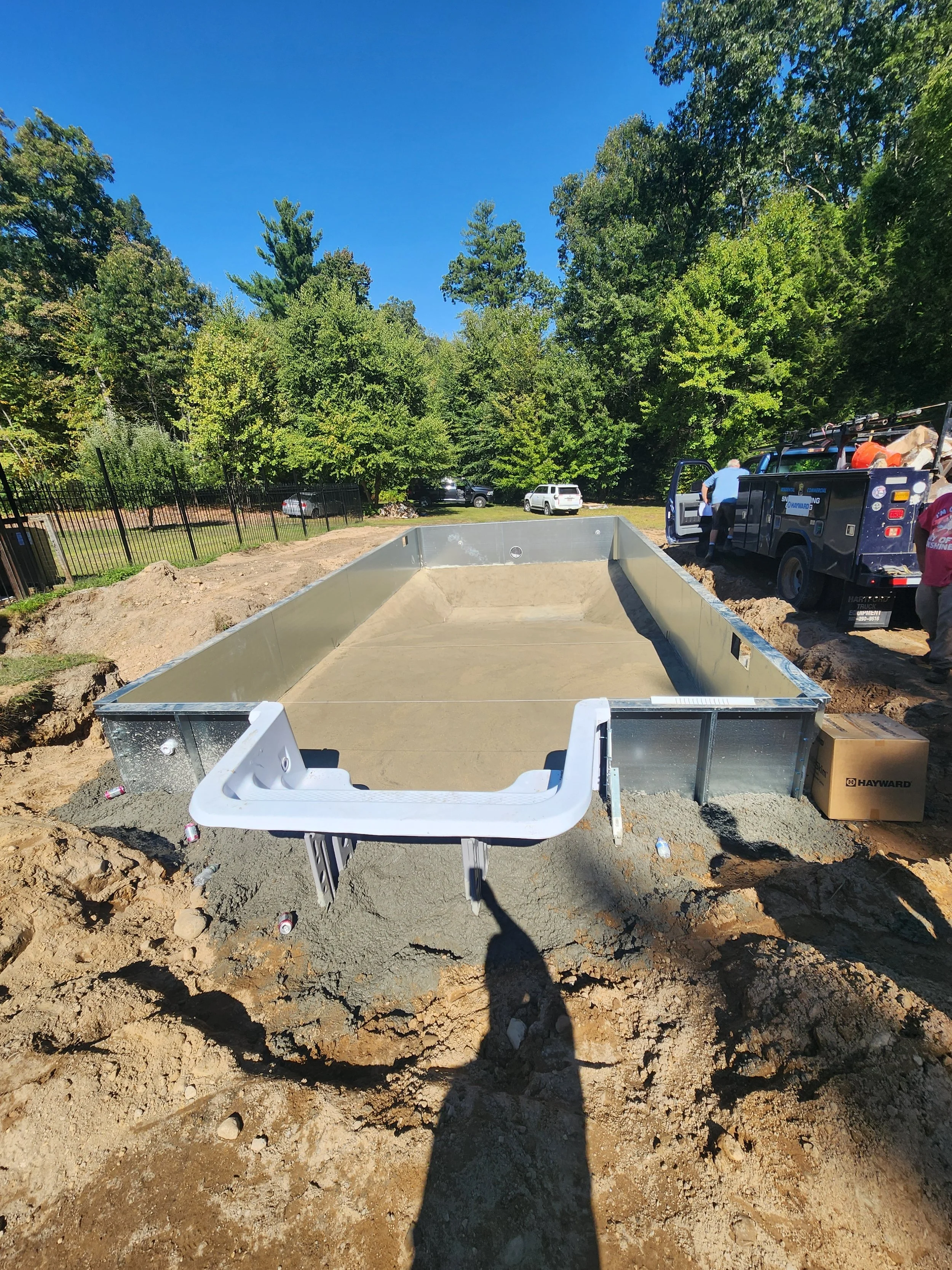 Construction site with dirt and a metal foundation frame, a utility sink, workers, parked cars, and trees under a clear blue sky.