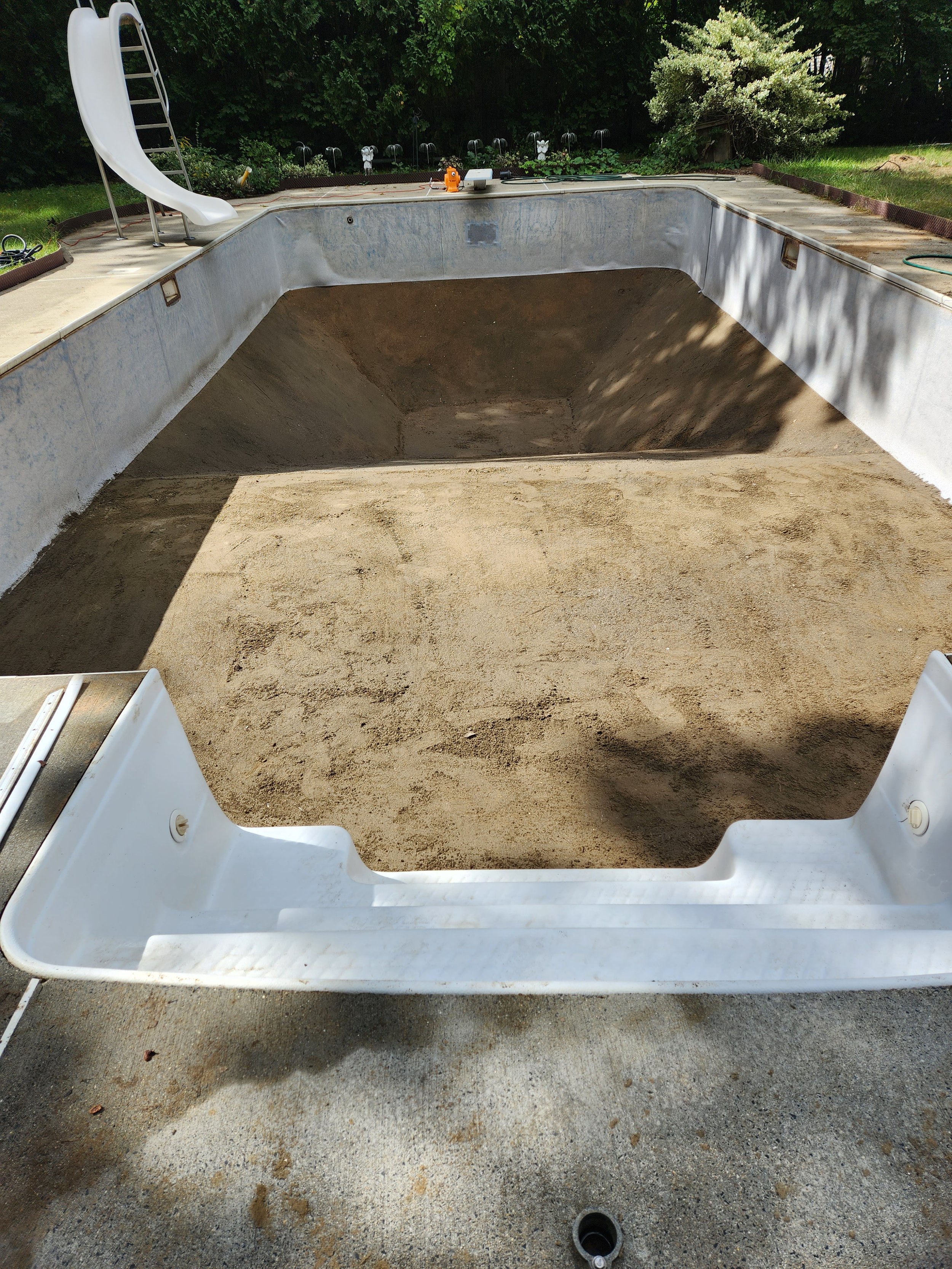 Empty swimming pool under renovation with dirt at the bottom, surrounded by a concrete deck and green garden foliage in the background.