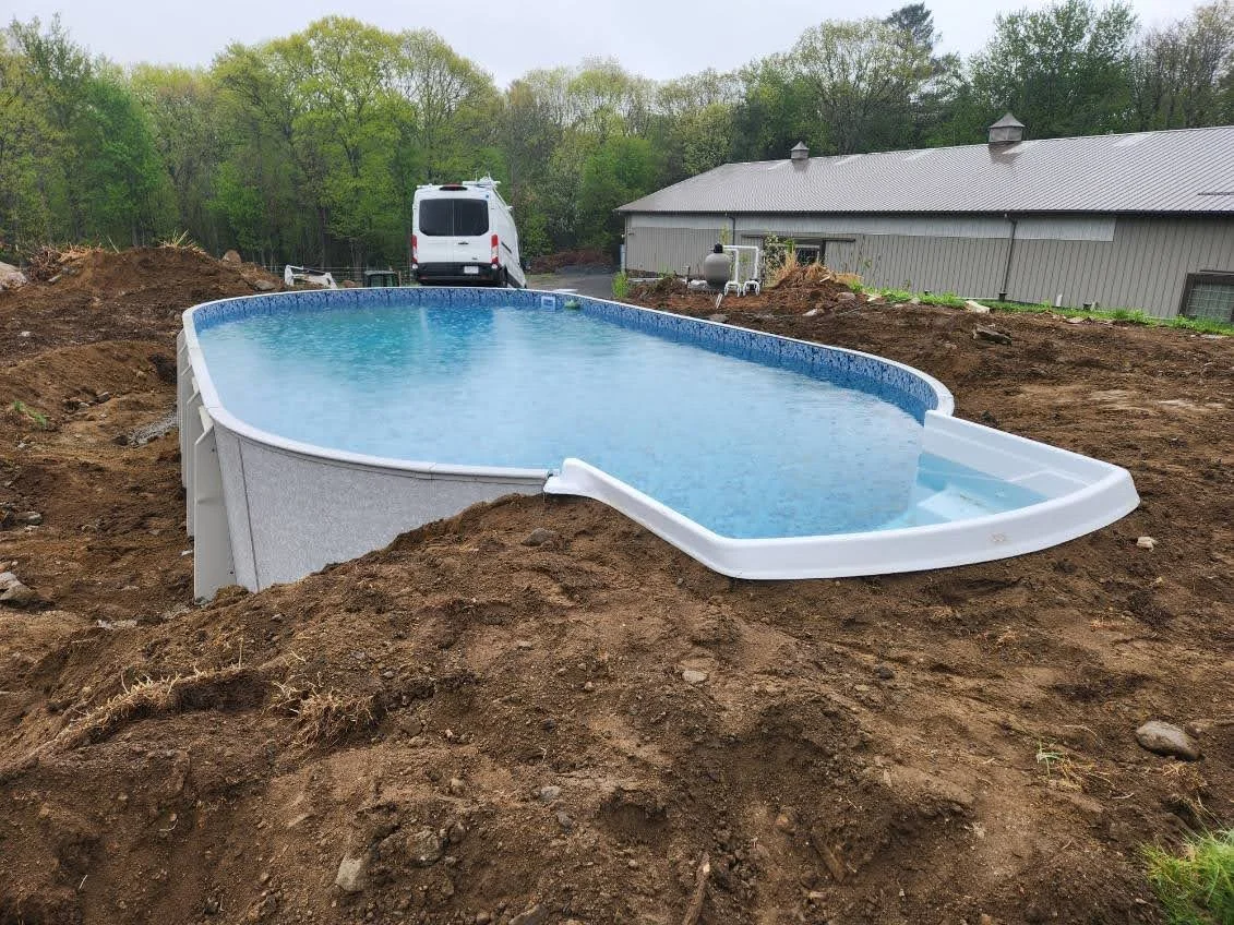 Above-ground rectangular swimming pool with water, partially installed in a backyard with dirt surrounding it, and a white van and building in the background.