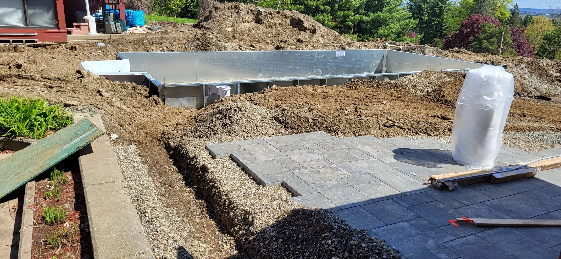 Construction site with a dugout area, a partially installed pond liner, and paving stones. There is a garden bed with green plants in the foreground, and trees and rolling hills in the background.