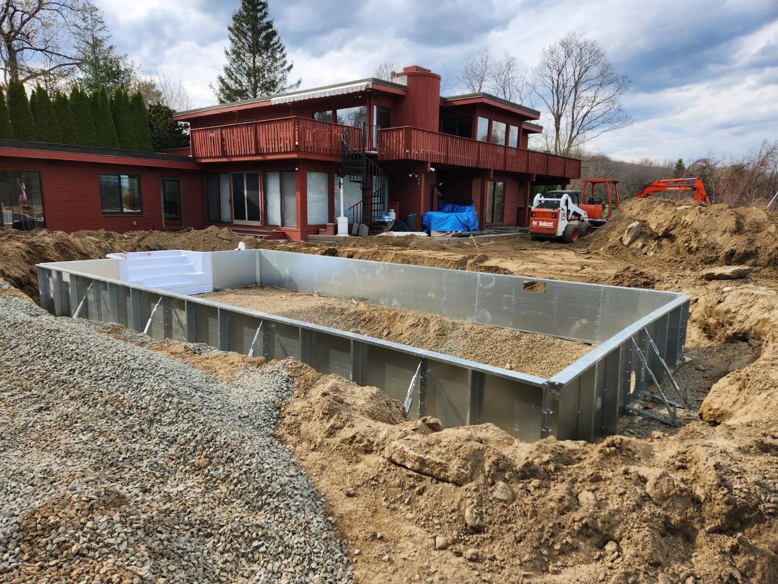 Under construction backyard pool with metal frame, concrete foundation, and construction equipment in a residential backyard.
