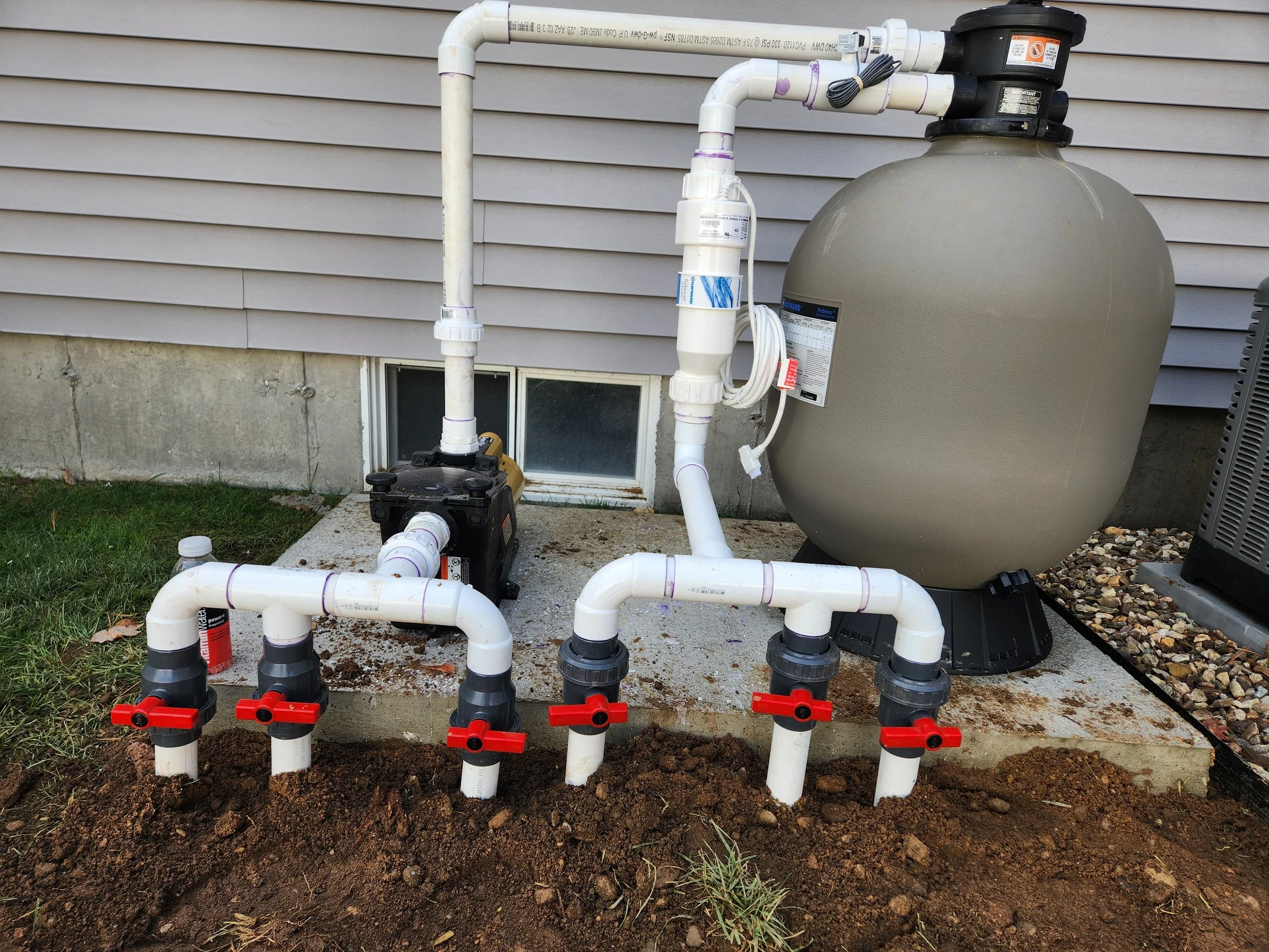 An outdoor sump pump system with white PVC pipes, a large beige tank, and black and red valves, installed against the house's foundation on a concrete slab with soil in the foreground.