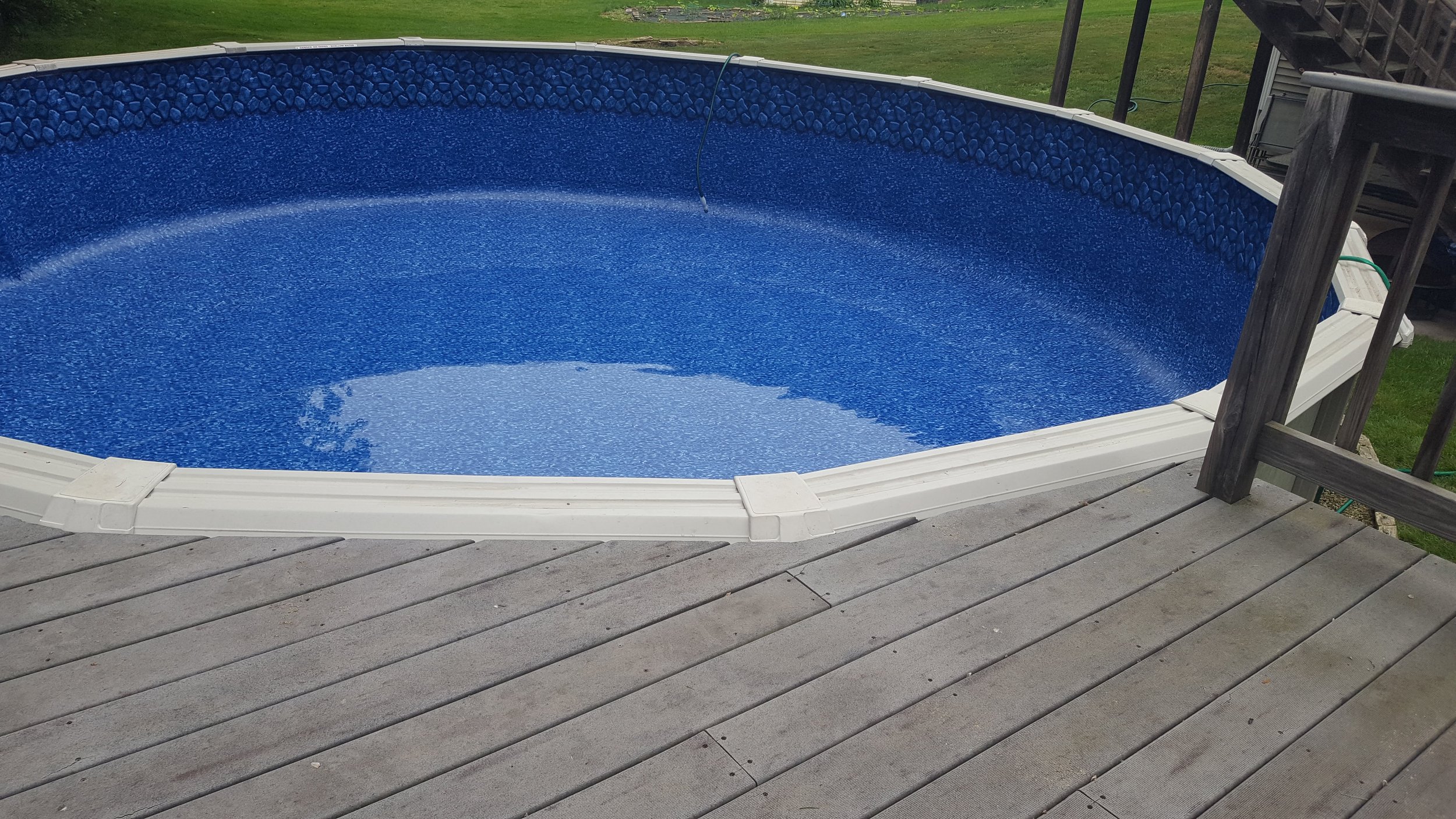 Above-ground swimming pool with a blue interior, partially filled with water, situated on a wooden deck in a backyard with green grass.
