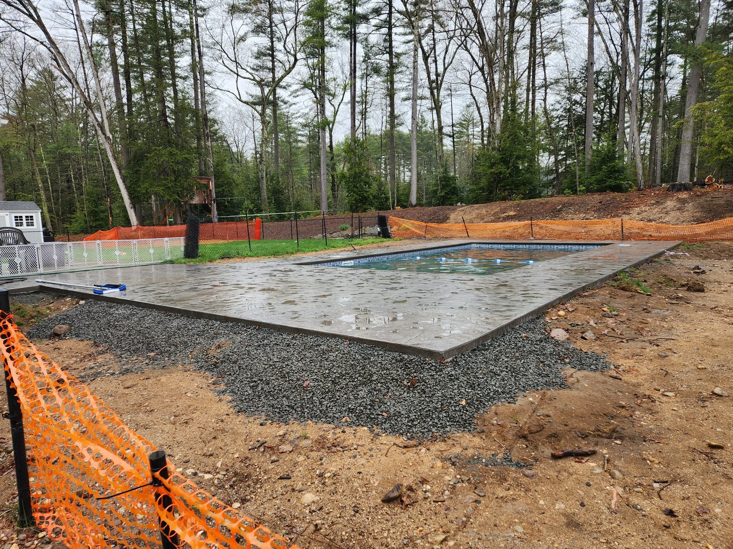 A backyard with a swimming pool under construction, surrounded by orange safety fencing, with a wooded area in the background and construction materials on the ground.