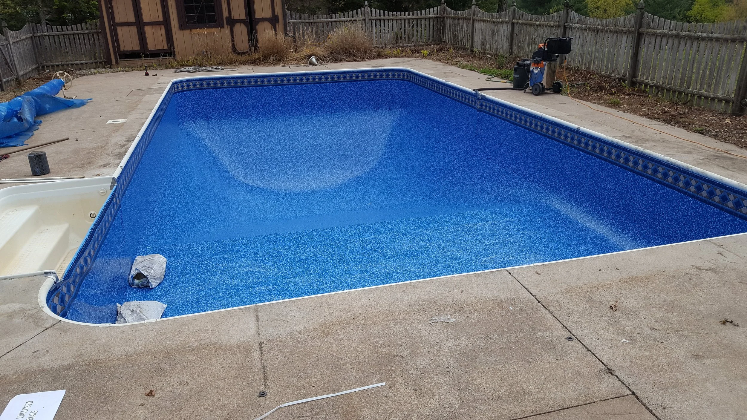 An empty swimming pool with a blue patterned interior on a concrete patio, surrounded by a wooden fence, with construction and cleaning equipment nearby.