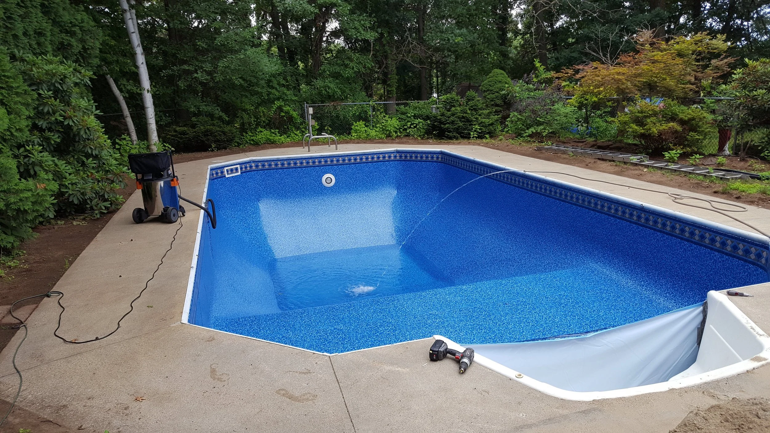 An in-ground swimming pool with a blue interior, surrounded by a concrete deck, in a backyard with trees and bushes. The pool appears to be empty, and there are tools and equipment nearby, suggesting ongoing maintenance or installation.
