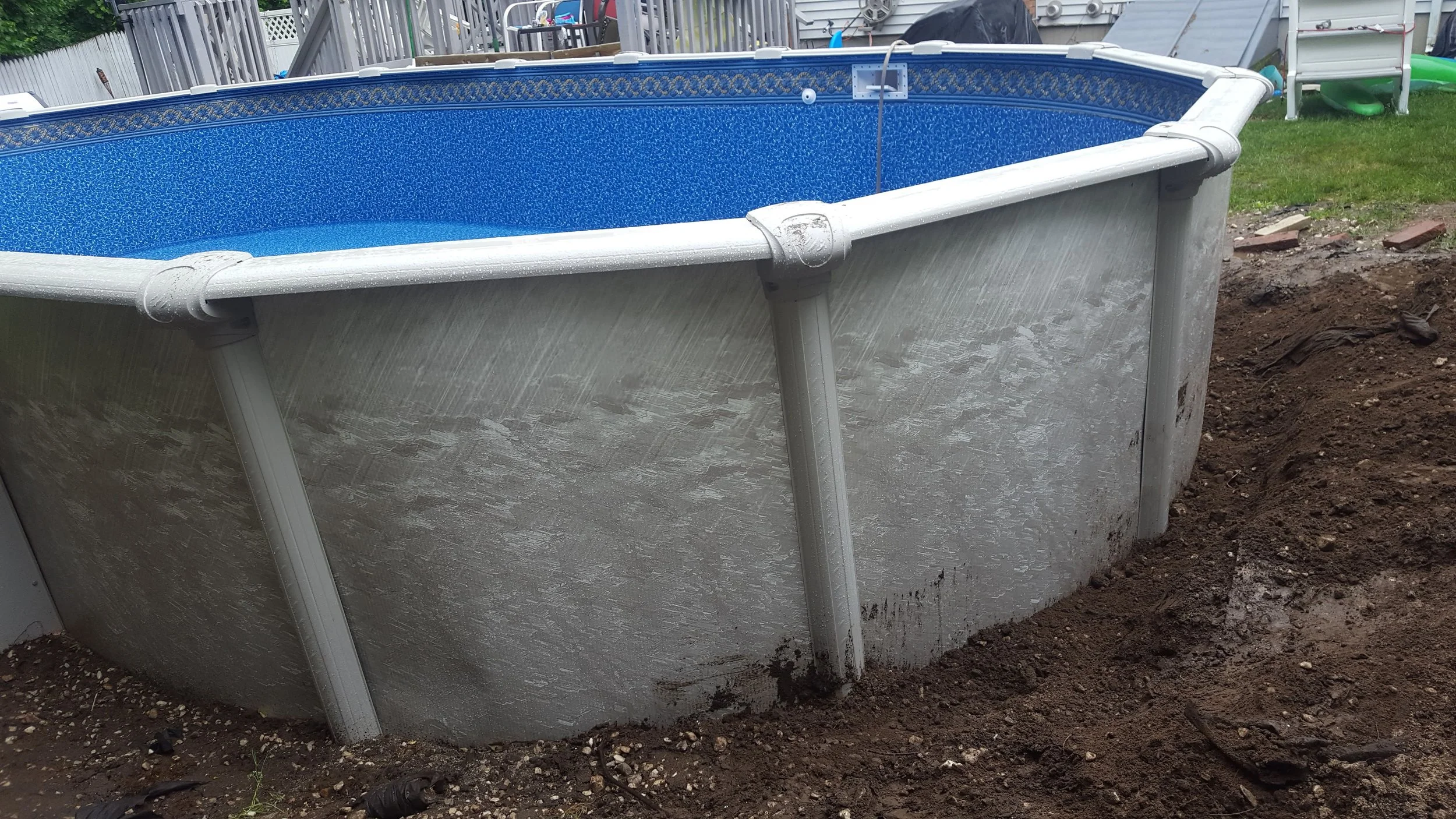 Above-ground swimming pool with a blue interior, partially filled with water, situated on a dirt yard in a backyard.