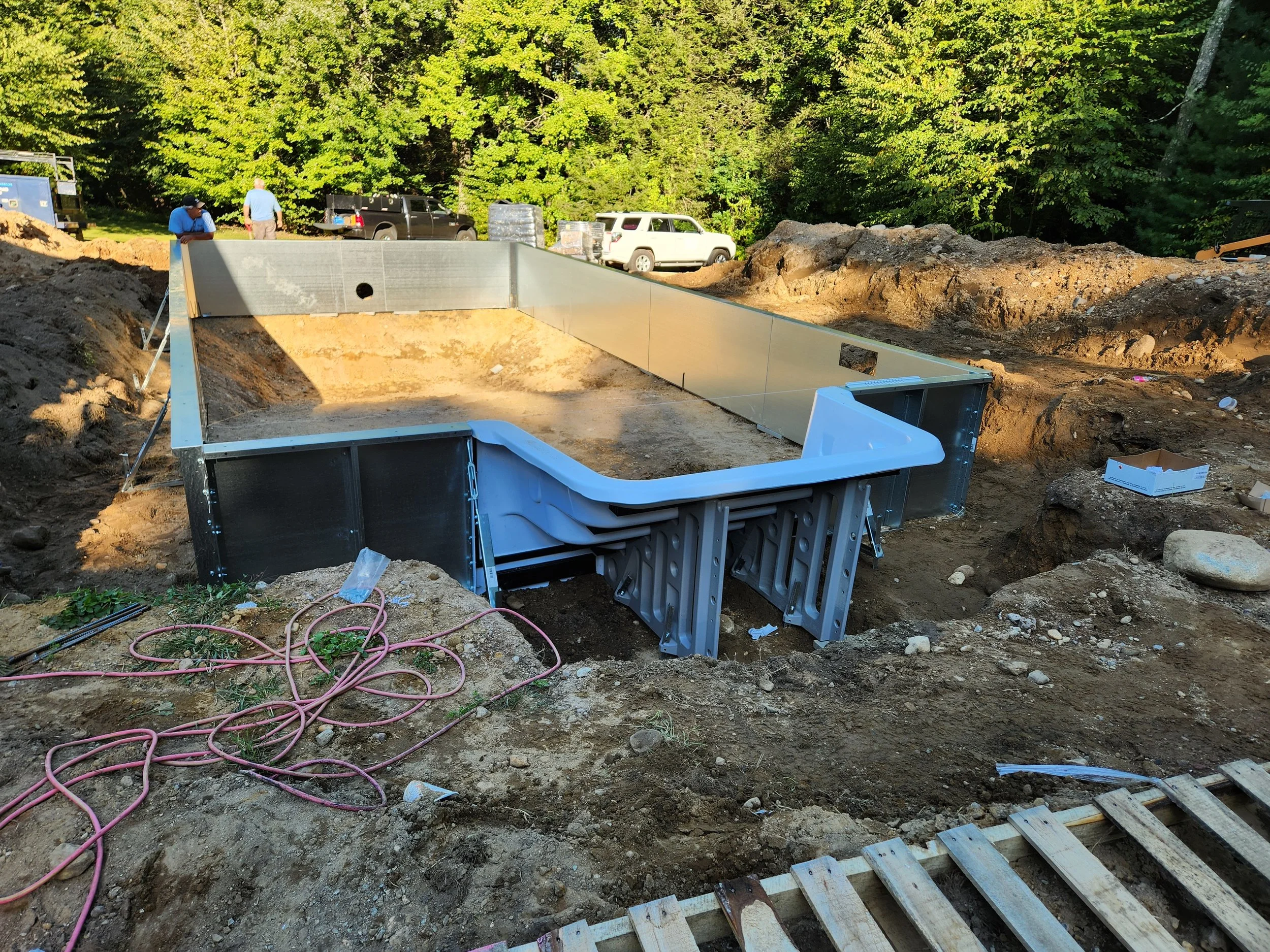Construction site with dug-out hole, metal framing, construction tools, on uneven dirt terrain surrounded by trees, vehicles, and construction equipment.