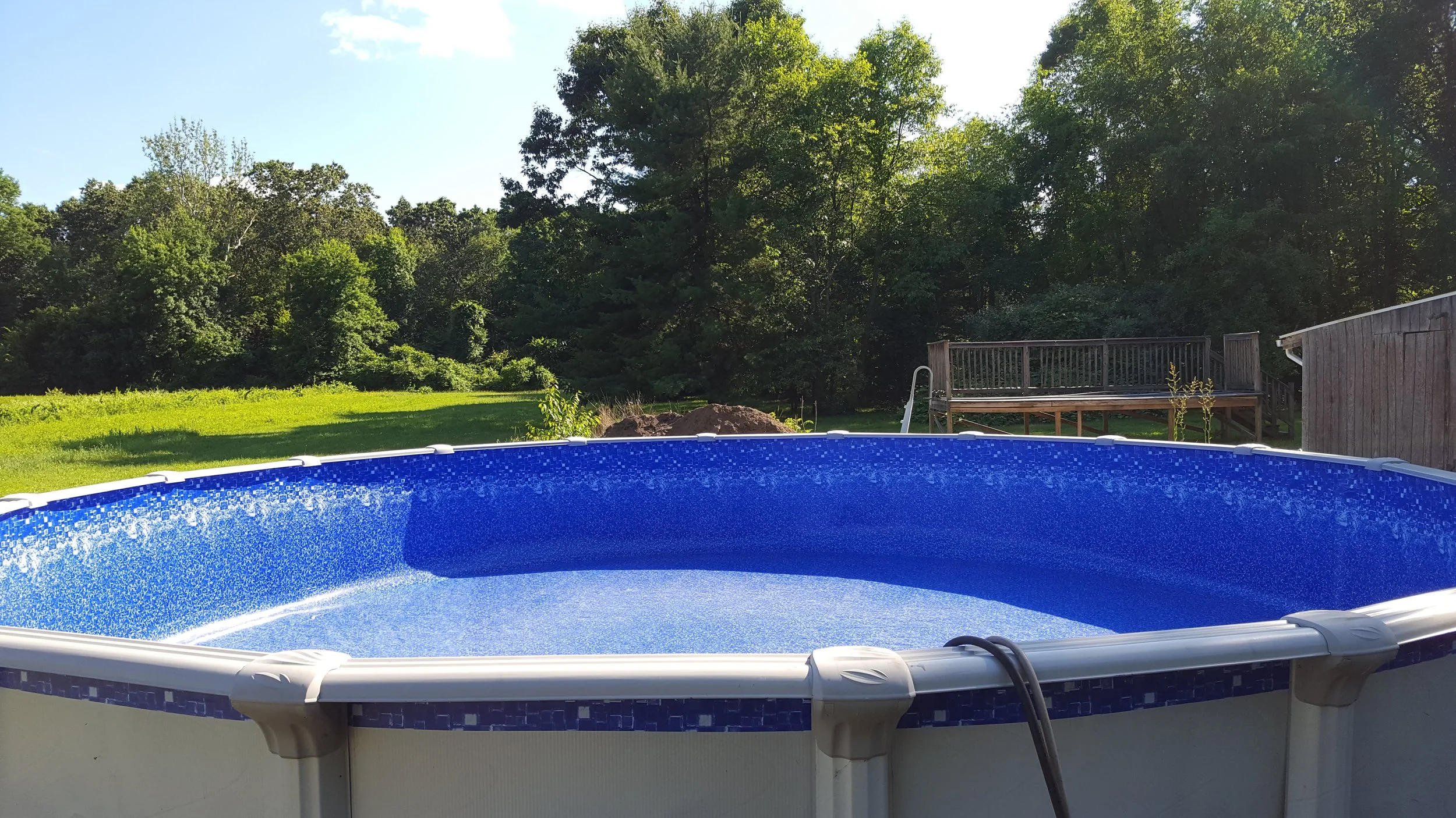 Above ground blue swimming pool in backyard with trees, green grass, wooden deck, and blue sky with clouds.