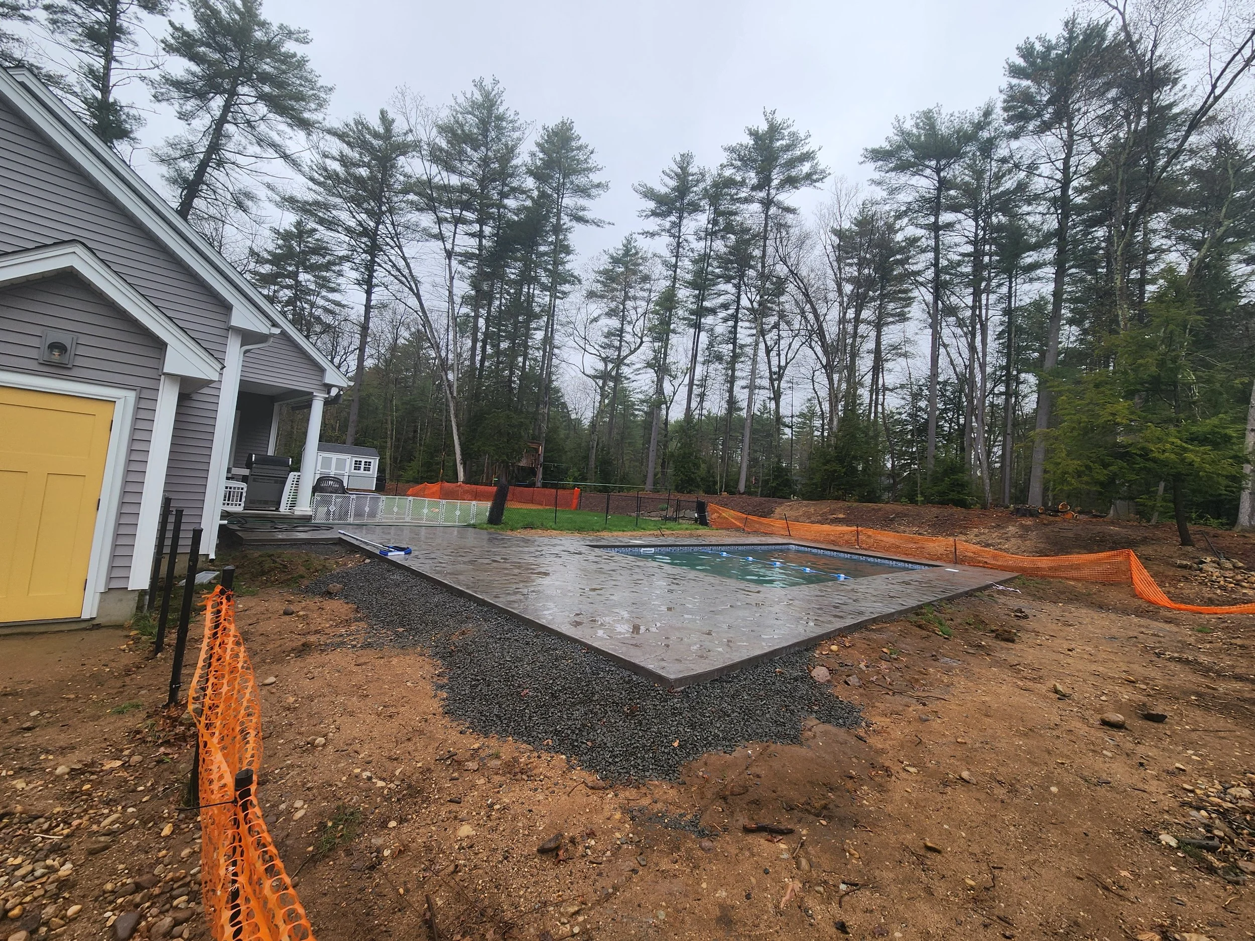 A backyard area with a newly poured concrete slab for a pool with orange safety fencing around it, adjacent to a house with gray siding and a yellow garage door, and surrounded by tall trees on a cloudy day.