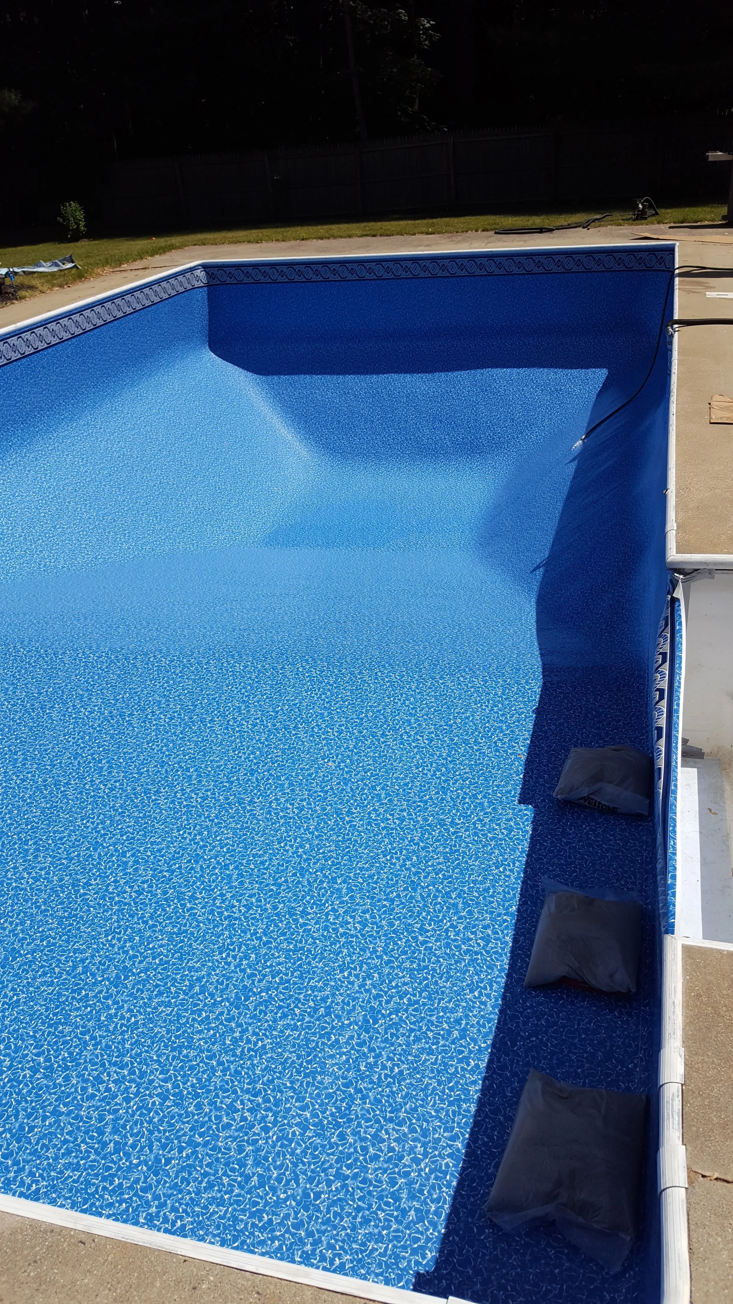 Empty swimming pool with a blue interior in a backyard. The pool is surrounded by a concrete deck and a black fence, with grass and trees visible in the background.