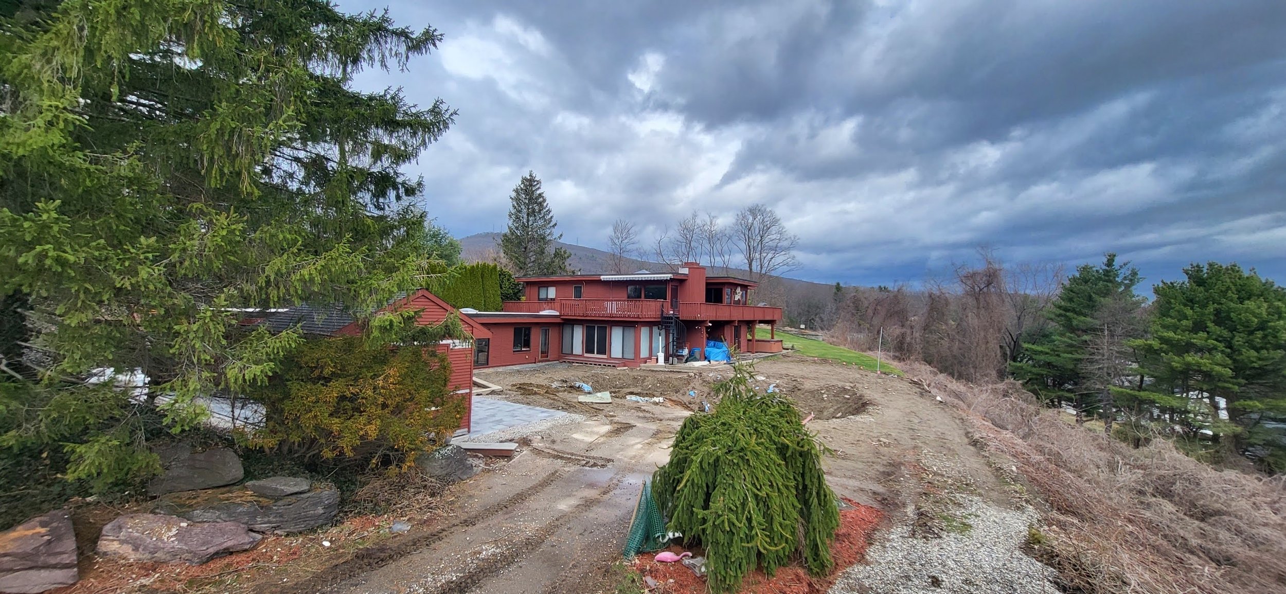 A two-story red house under construction on a hillside with a dirt yard and surrounding trees. Overcast sky with dark clouds.