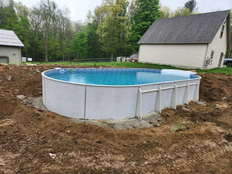 Inground above-ground swimming pool under construction in a backyard, with dirt around it and a house in the background.