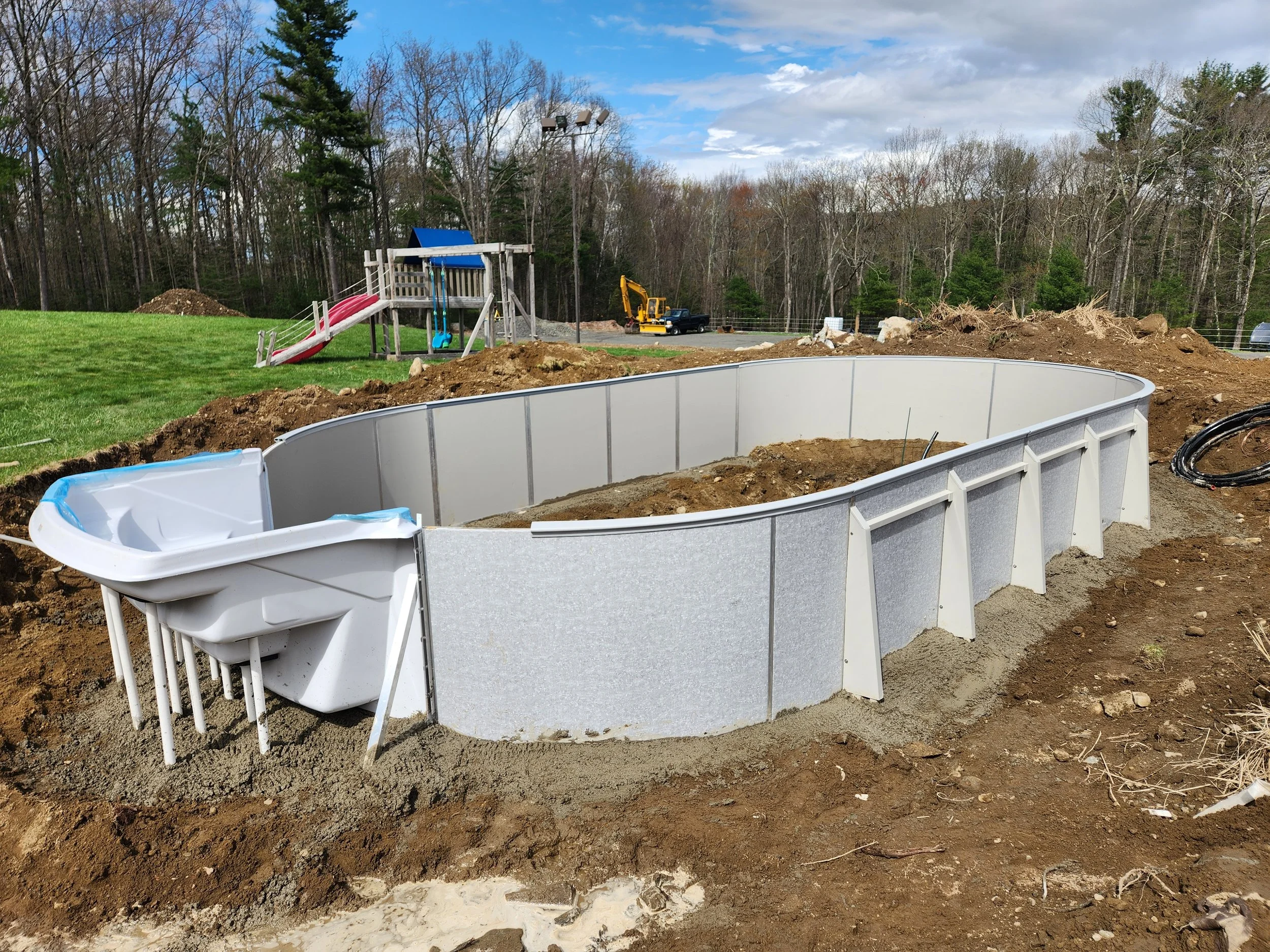 A backyard swimming pool under construction with dirt around it, a playset with a slide and swings, a small shed, and construction equipment, with a backdrop of trees and a partly cloudy sky.