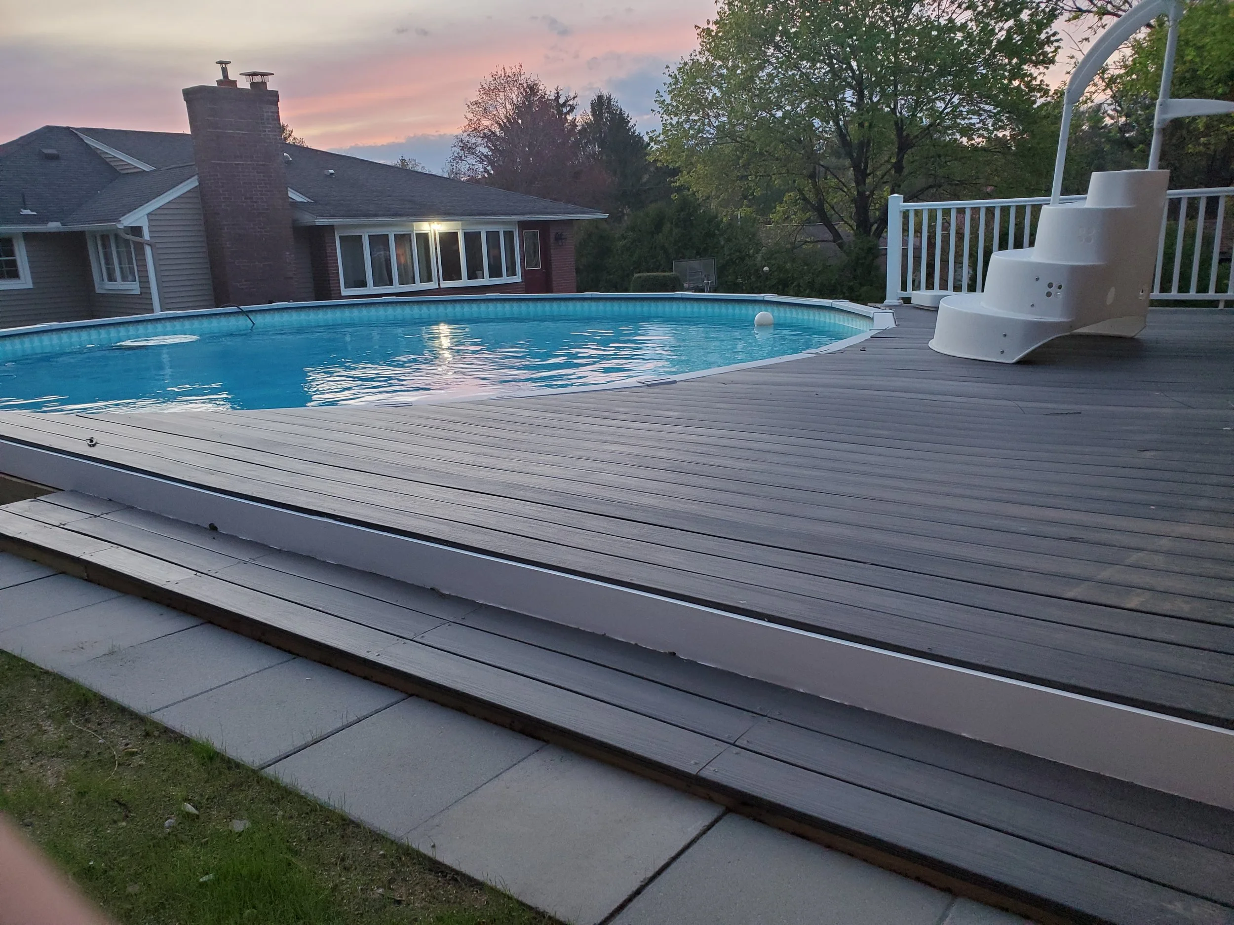 A backyard with an above-ground swimming pool on a wooden deck, surrounded by a white railing, with trees and a house in the background at dusk.