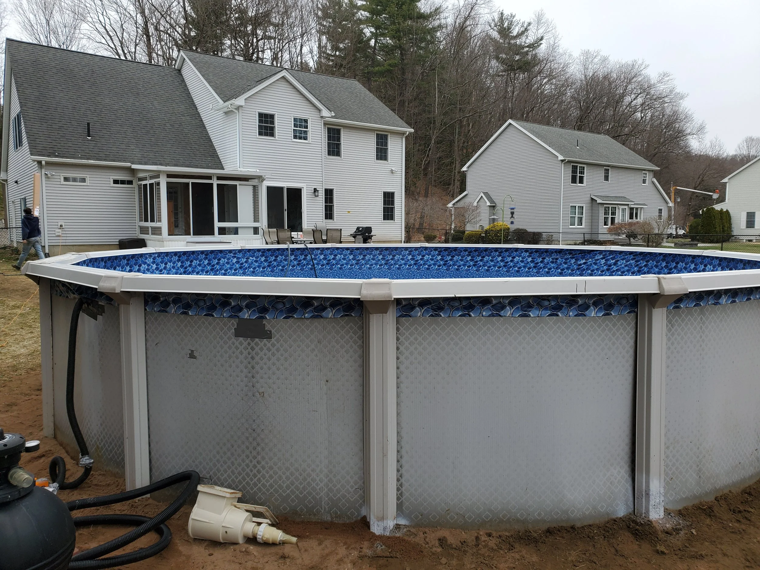 An above-ground swimming pool being installed in a backyard, with the pool partially assembled and tools scattered nearby, two houses and a wooded hill in the background.