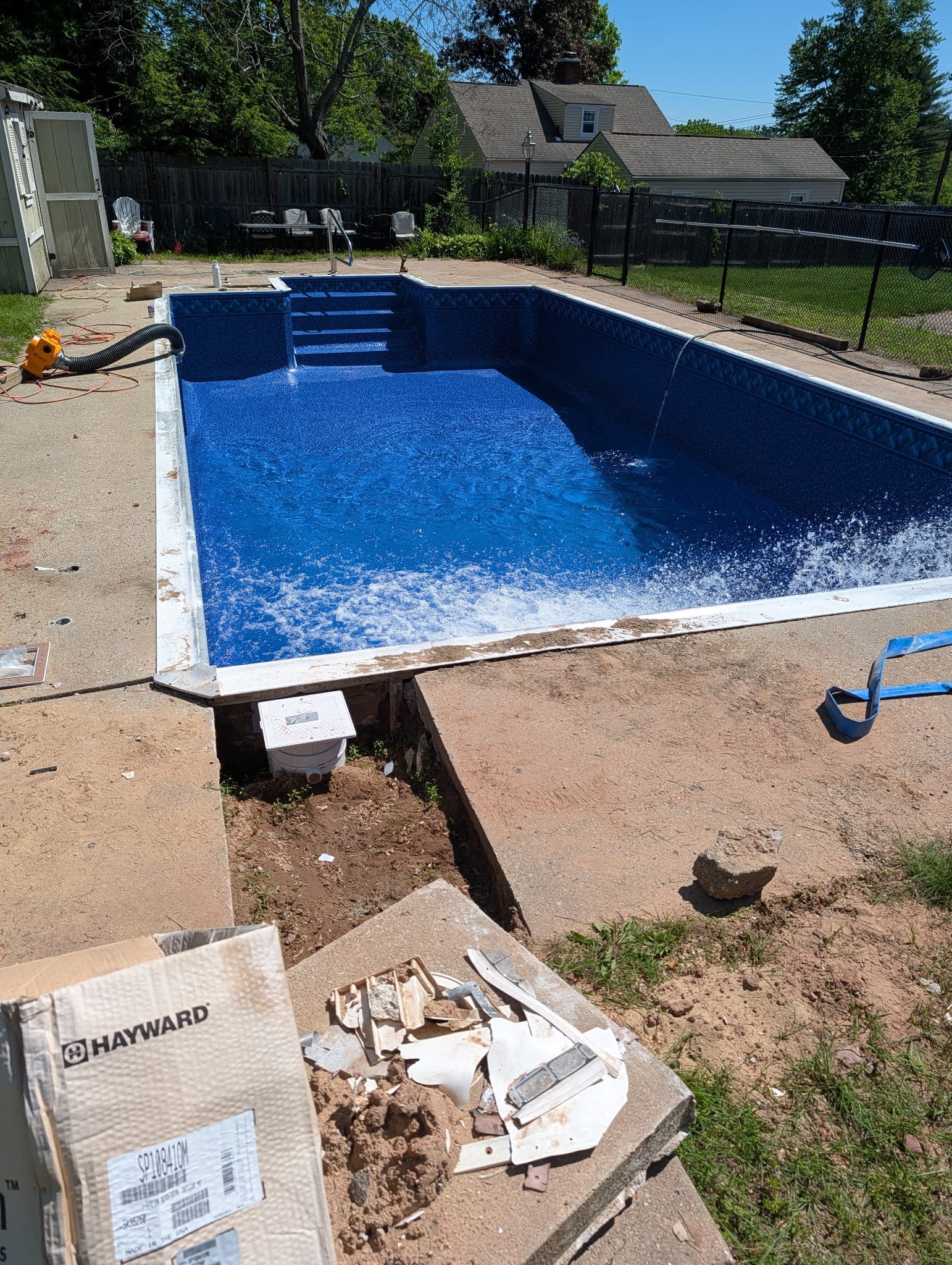 A backyard with a swimming pool under construction, surrounded by a black fence, with building materials, tools, and debris on the ground. In the background, there are trees, a house, and a shed.