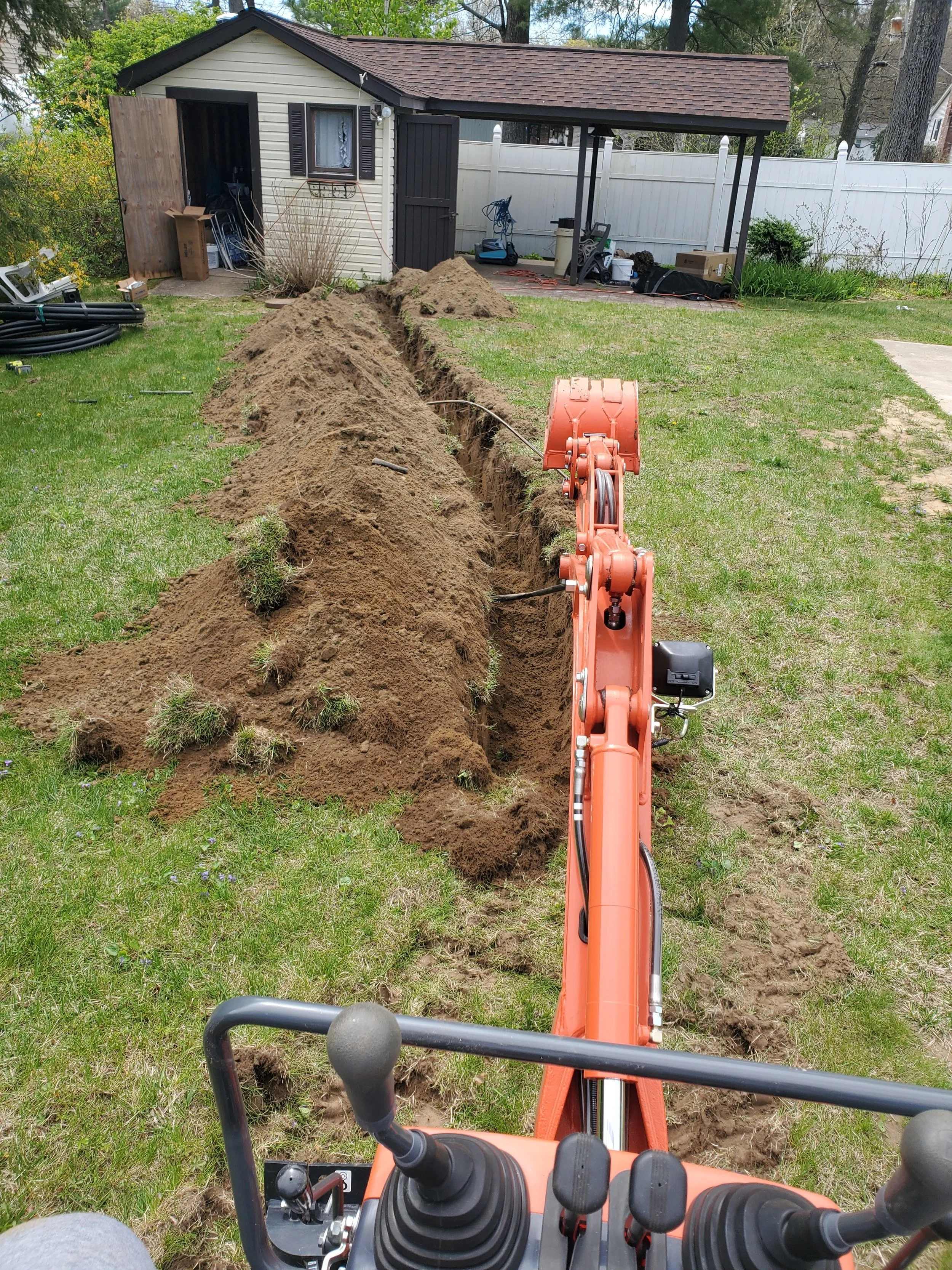 View from a construction vehicle, showing a freshly dug trench in a backyard, with a small shed and various tools and equipment around.
