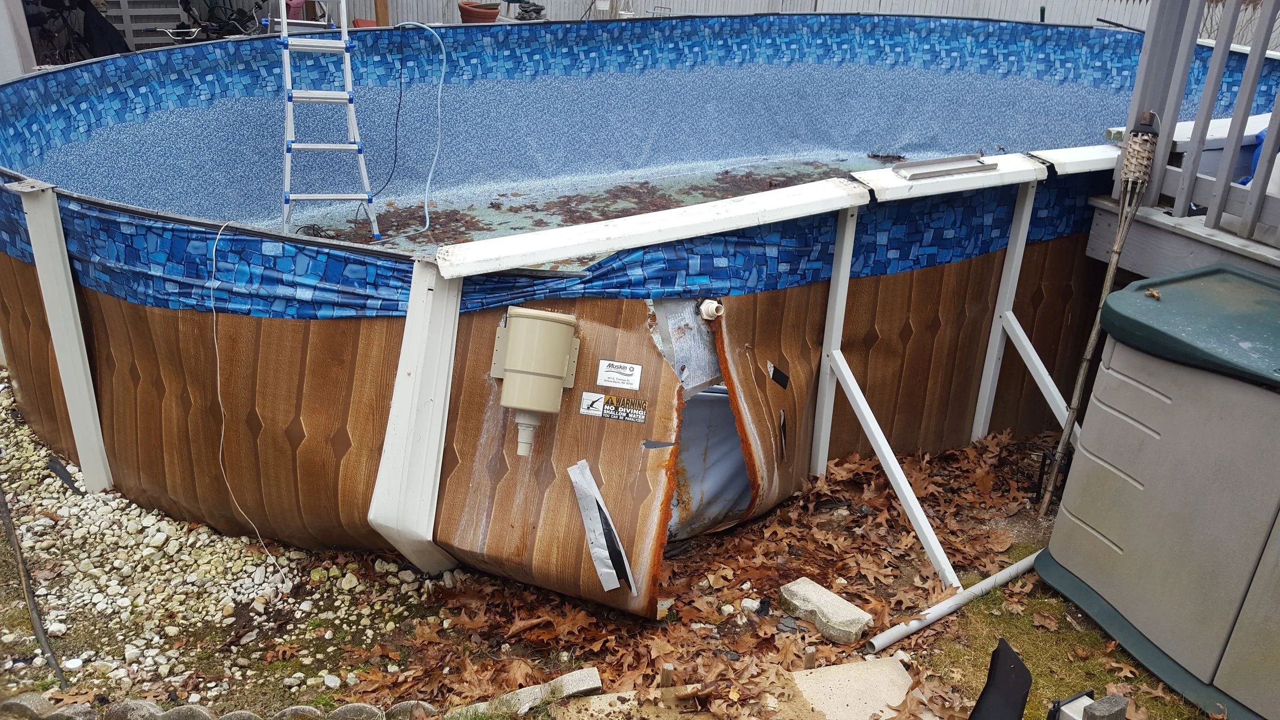 In-ground swimming pool with damaged and broken liner, debris, leaves, and pool equipment around it, with step ladder inside.