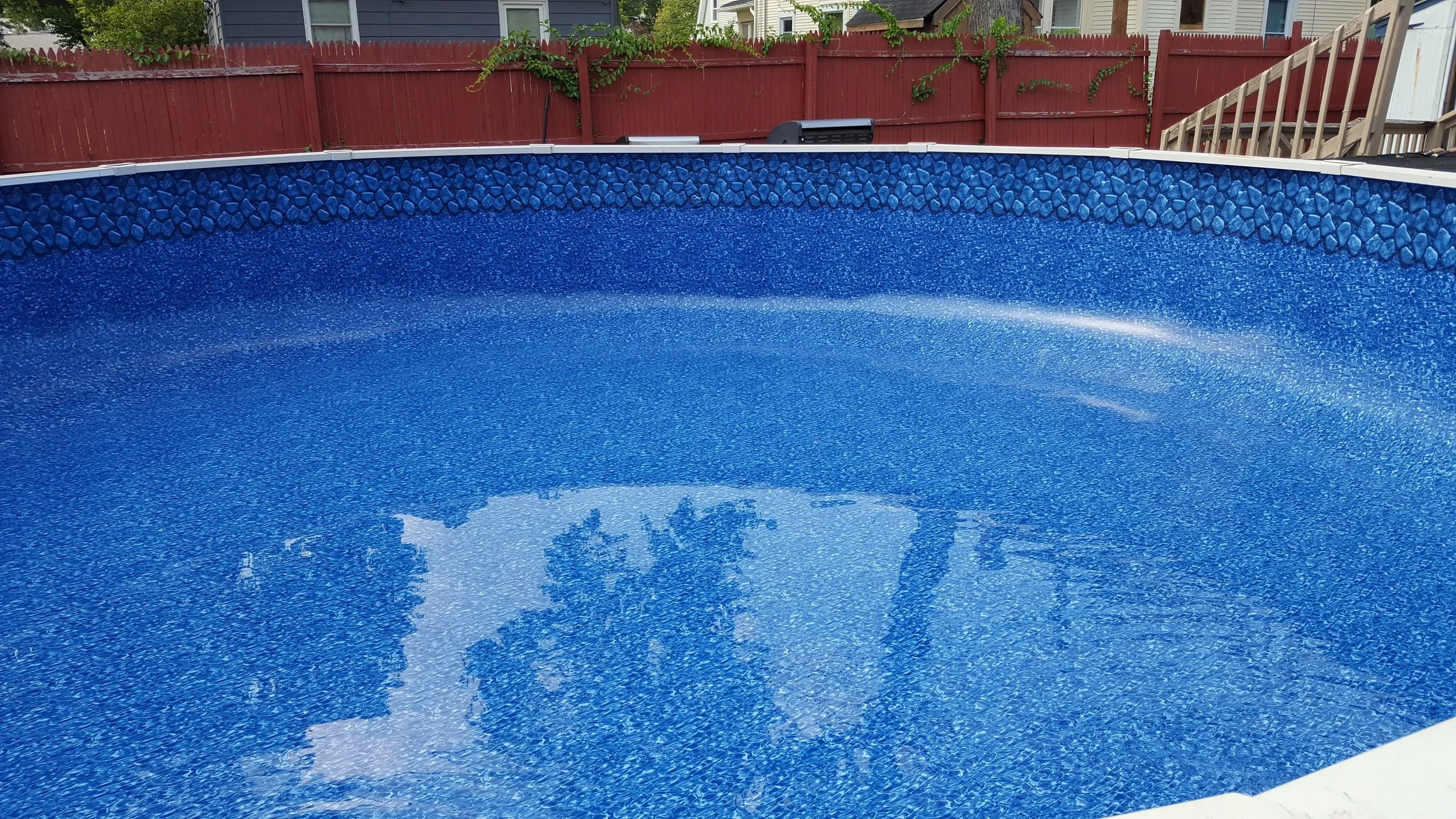 An empty above-ground swimming pool with a blue interior, surrounded by a red wooden fence, in a backyard with neighboring houses.