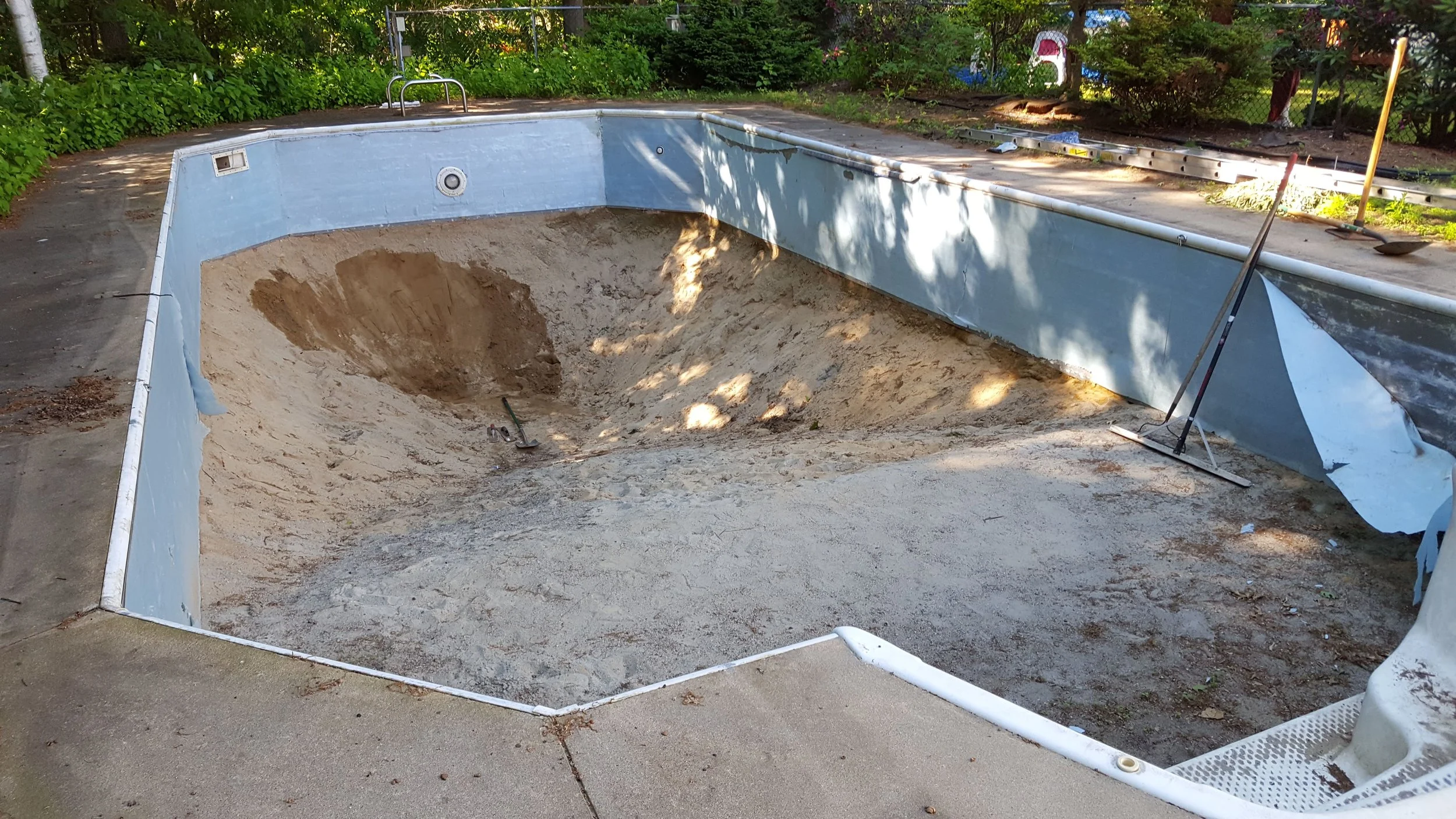 An empty backyard swimming pool undergoing renovation, with sand and dirt inside, and tools and supplies around the pool area.