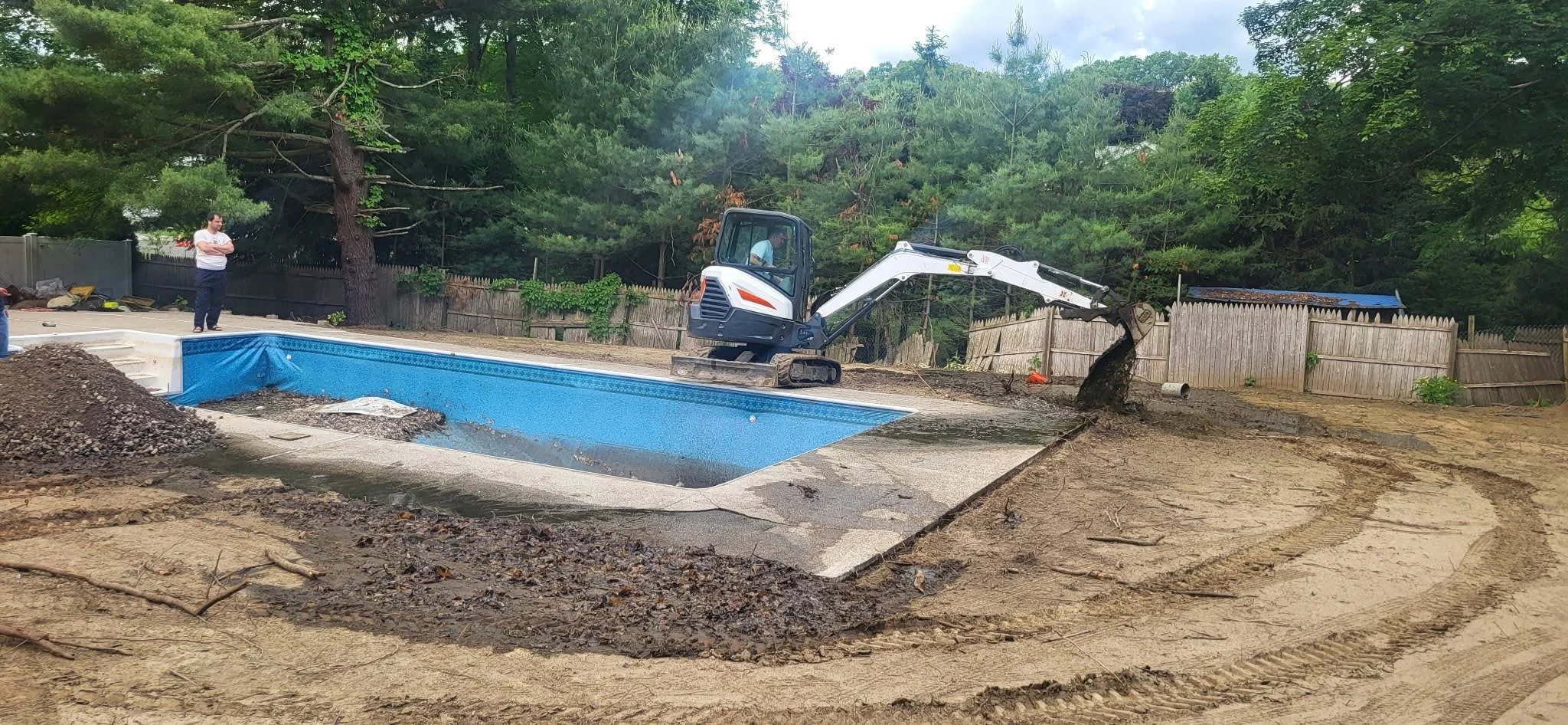 A backyard pool is being filled with dirt using a small excavator, with a person standing nearby observing.