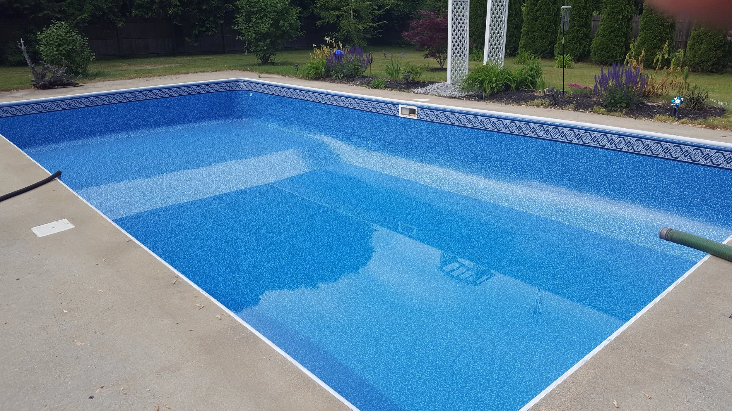 Empty in-ground swimming pool with blue mosaic tiles, surrounded by a concrete deck and landscaped garden with trees, bushes, and purple flowers.