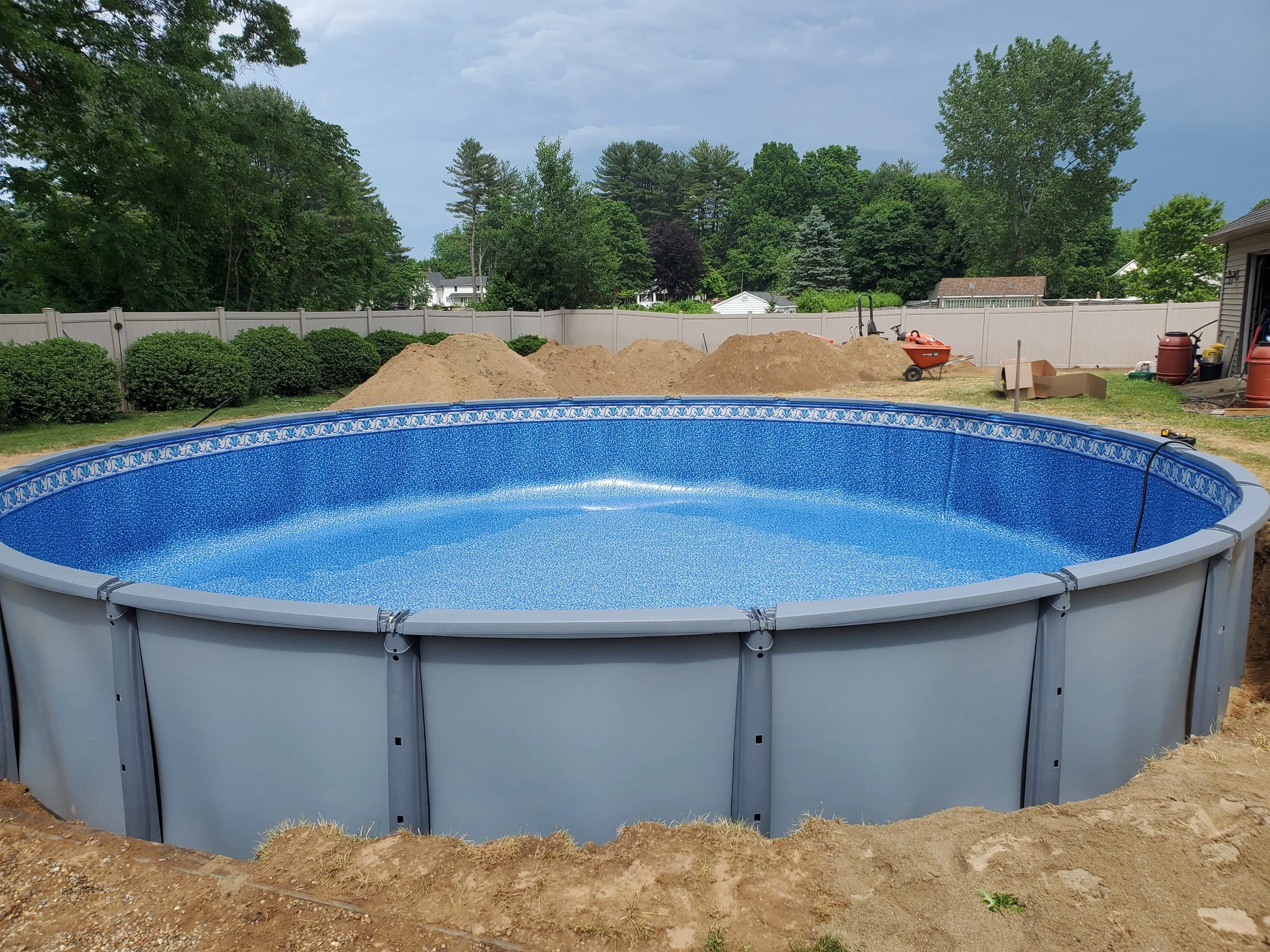 In-ground swimming pool in backyard with a dirt area next to it, a white fence in the background, and trees and houses beyond the fence.