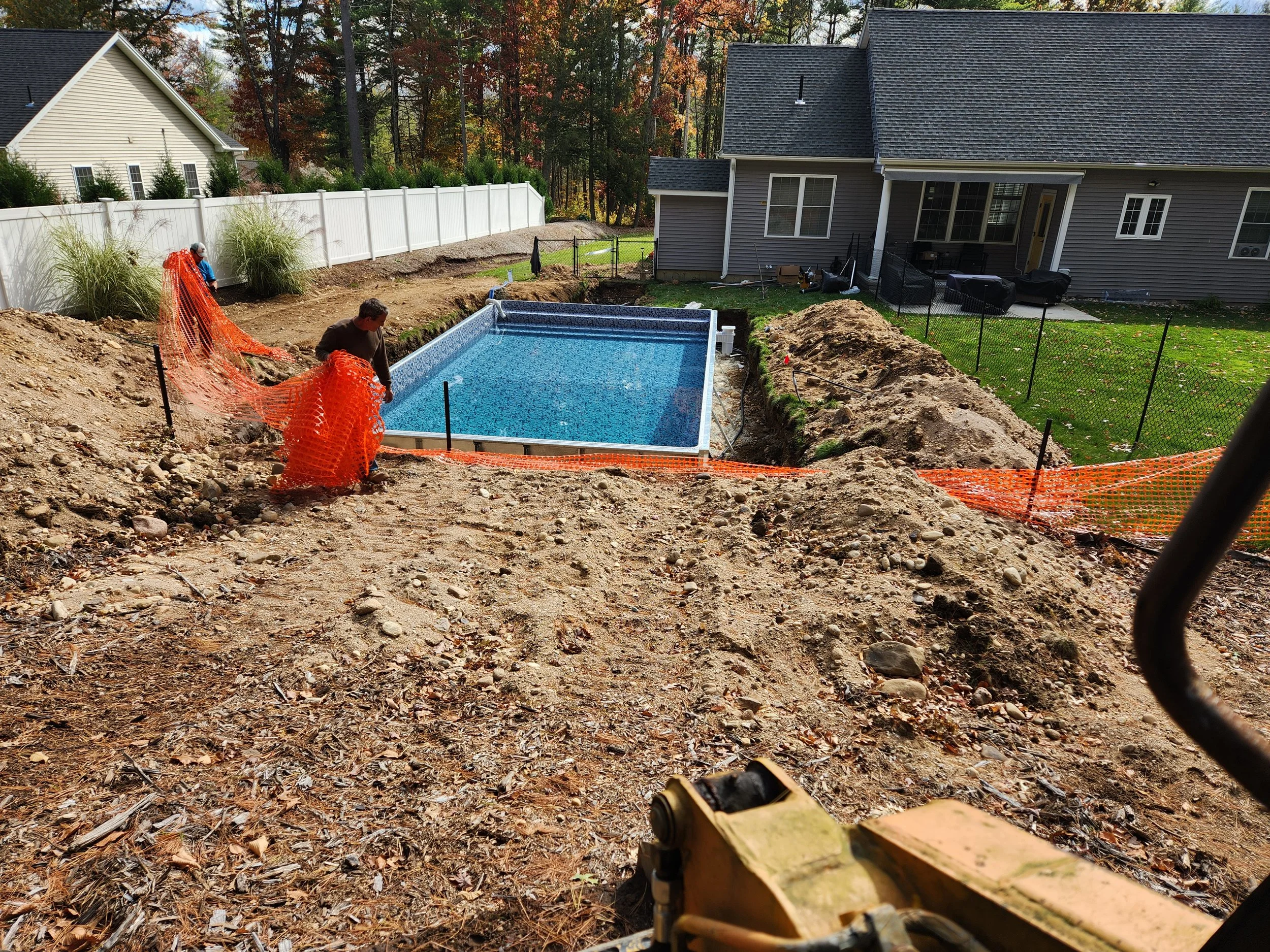 Backyard under construction featuring a rectangular in-ground swimming pool with a blue liner, surrounded by dirt piles and construction equipment, enclosed by a black mesh fence, with a gray house with a deck and outdoor furniture, and a white fence