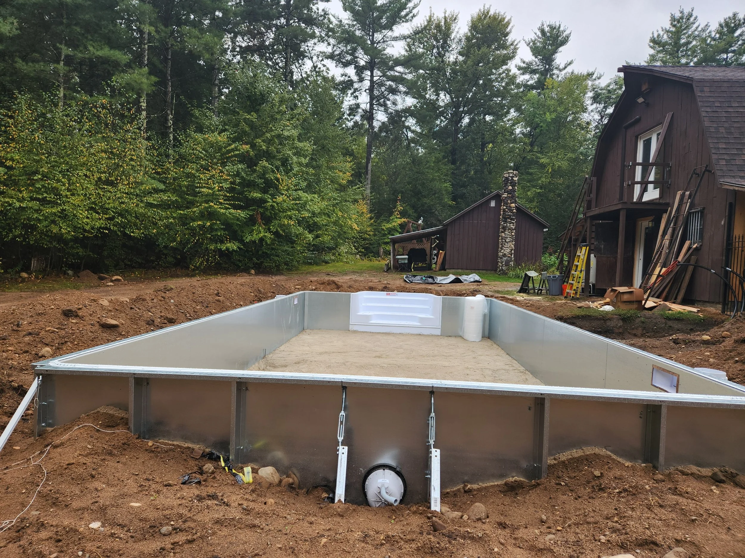 Construction site with a partially built swimming pool in the foreground, surrounded by dirt and construction materials. In the background, there are wooden houses and dense trees.