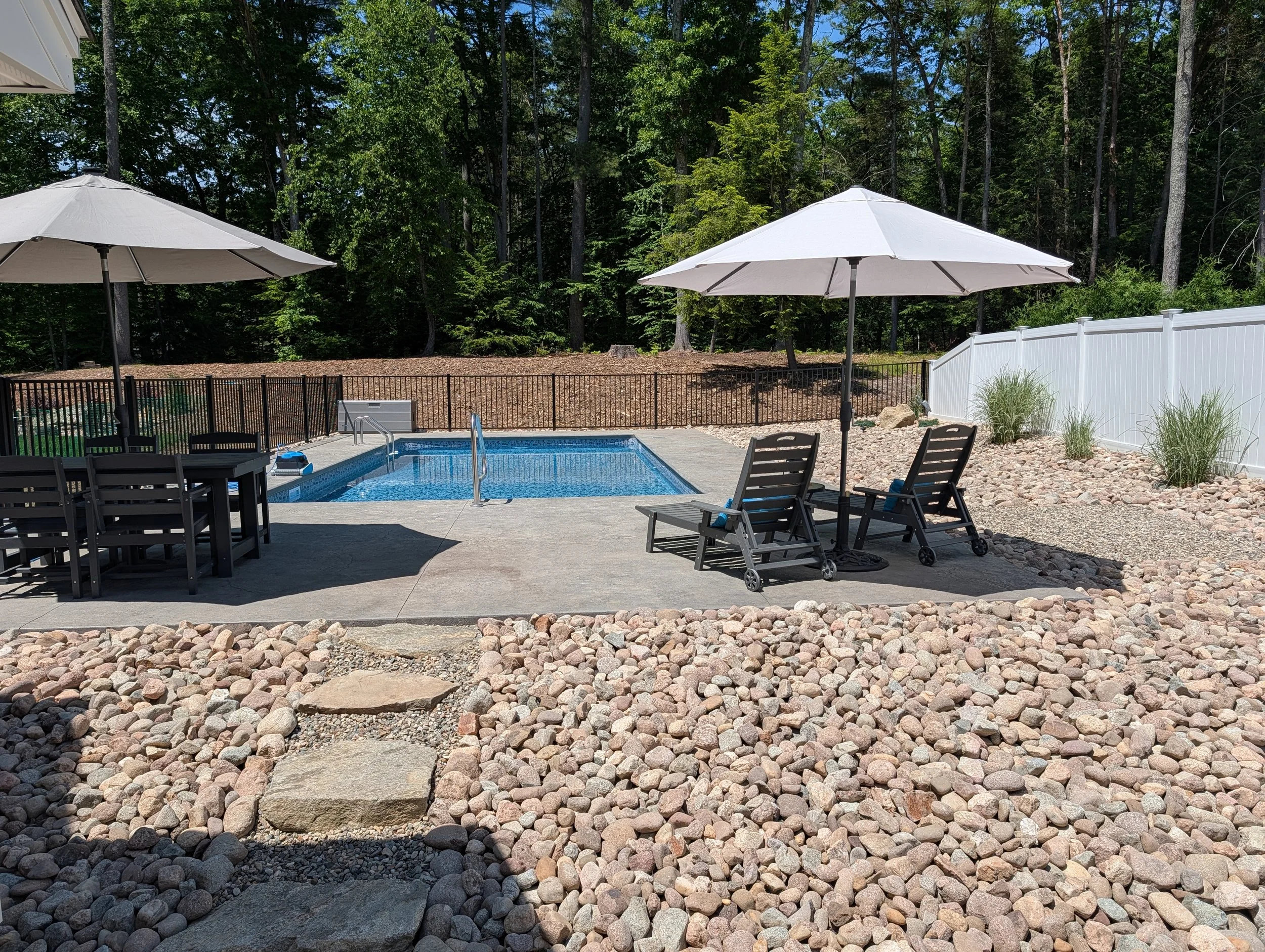 Backyard pool area with two white patio umbrellas, black chairs, and a privacy fence, surrounded by rocks and trees.