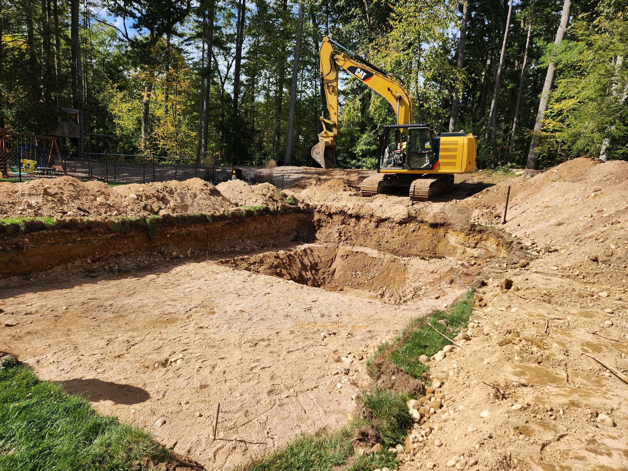 Construction site with an excavator digging a large excavation in the ground surrounded by trees.