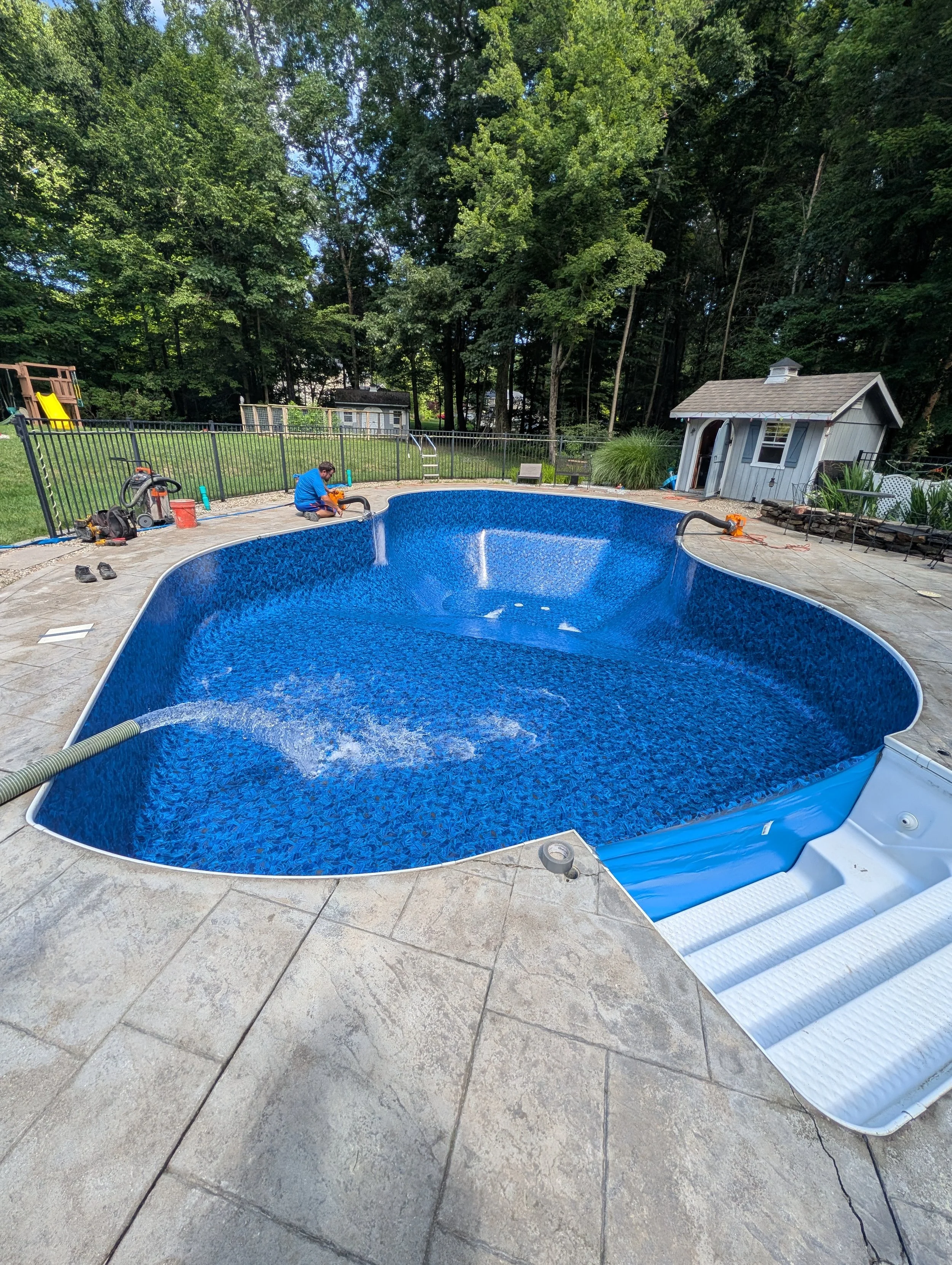 A backyard with a kidney-shaped in-ground swimming pool, one person working on the pool, a small shed, a fenced yard, a play set, and a surrounding forested area.