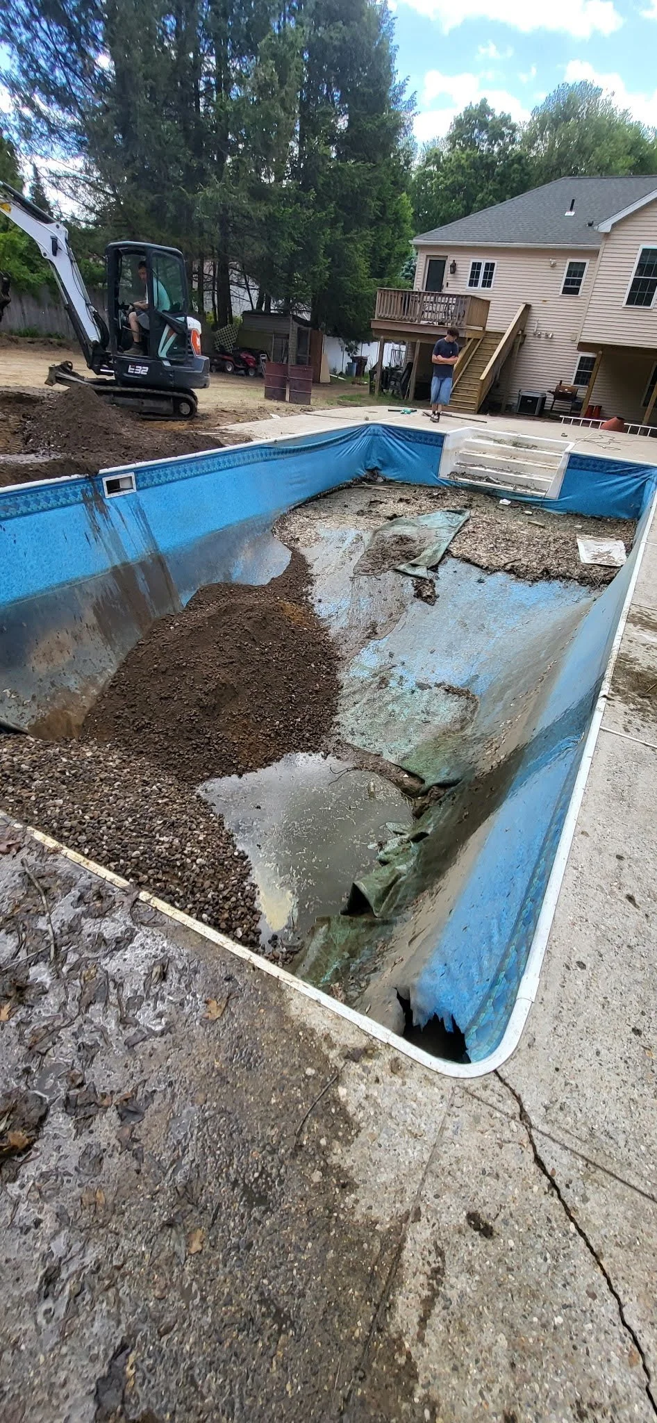 Empty and damaged swimming pool with debris and water, construction equipment, and two people in the background working on yard renovation.
