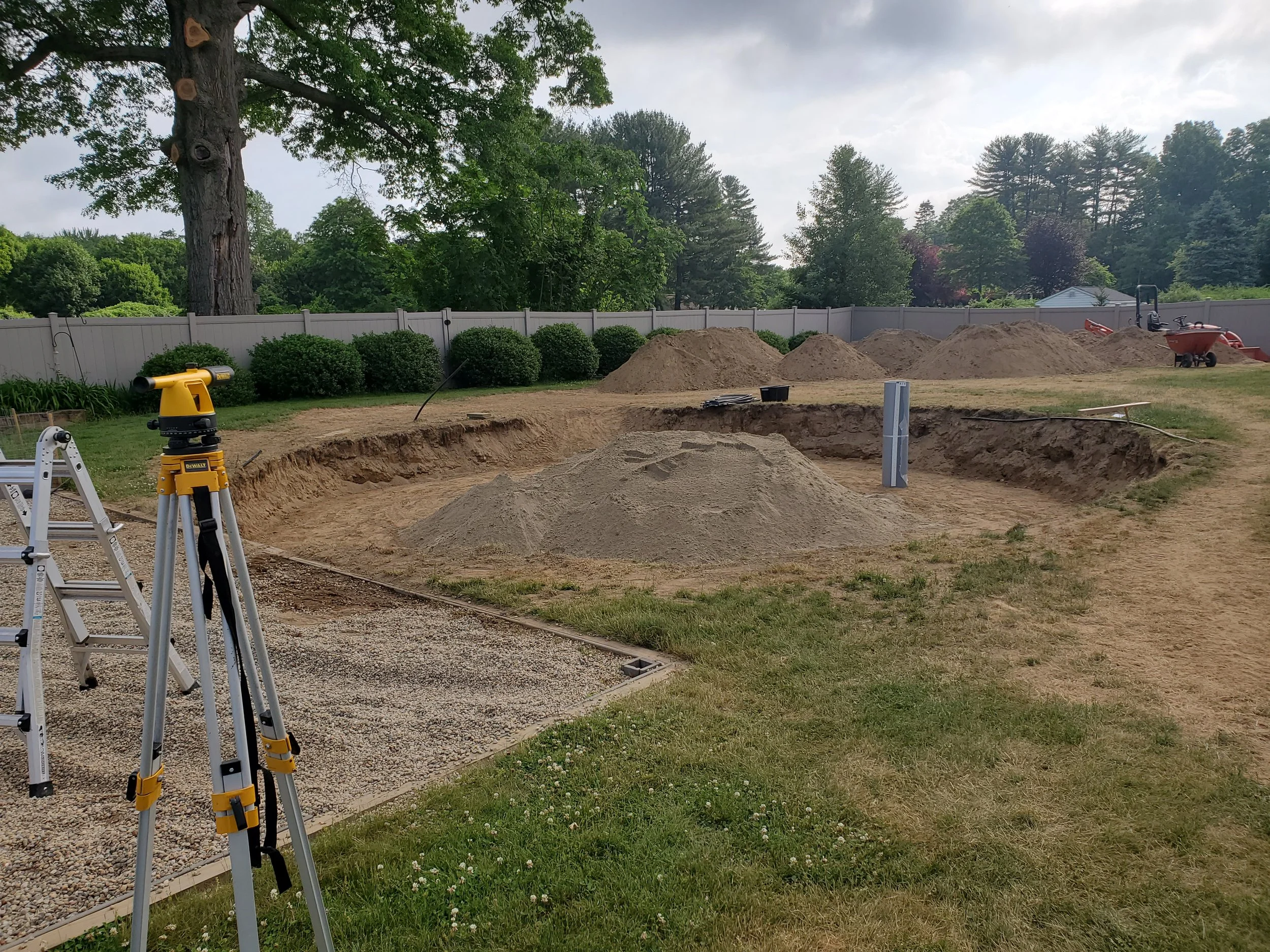 A backyard under construction with piles of dirt, construction tools, and equipment including a level, wheelbarrow, and surveying instrument. Trees and a white fence line the background.