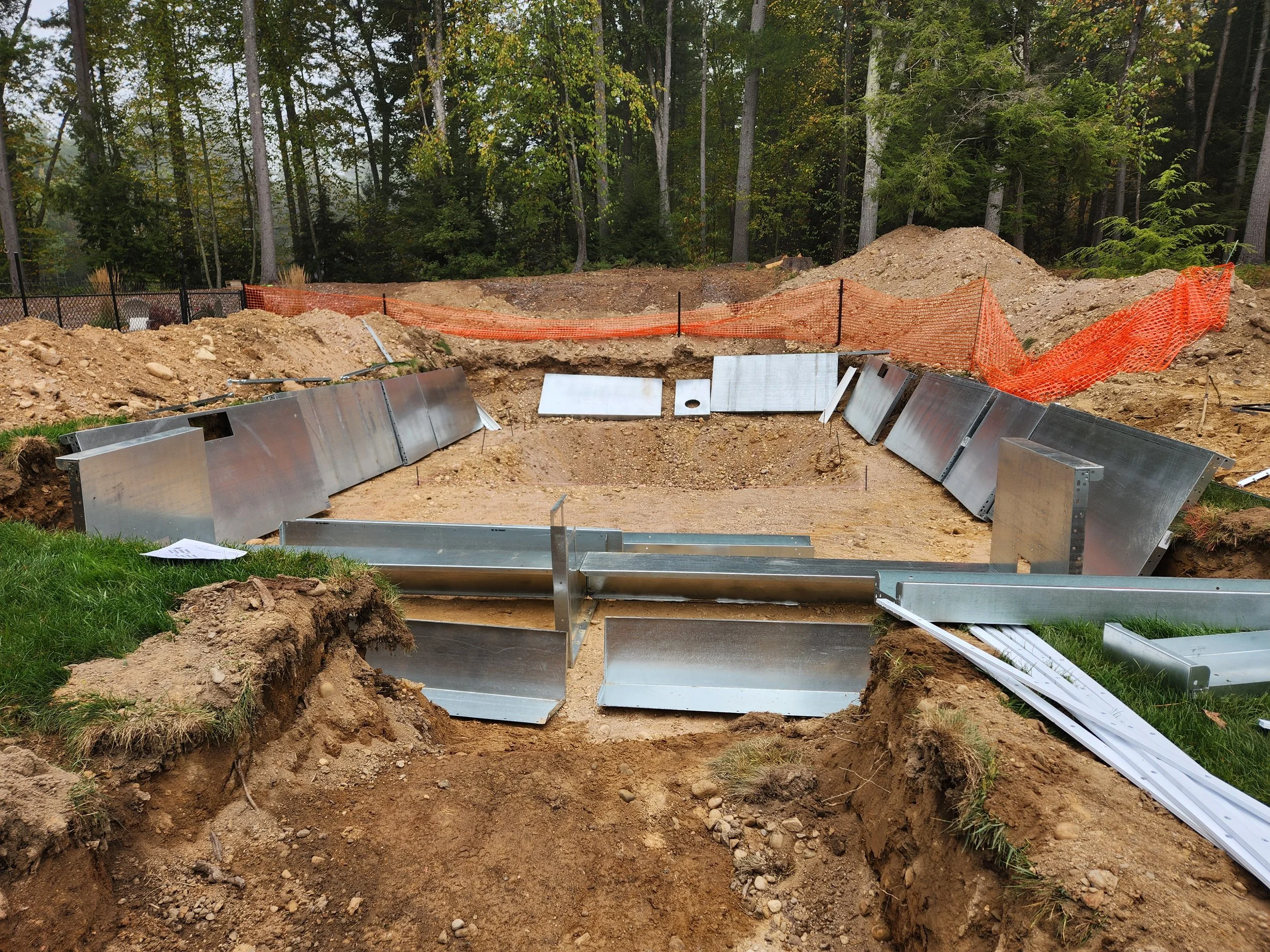 Construction site with excavated area, metal barriers, orange safety netting, and a forested background.